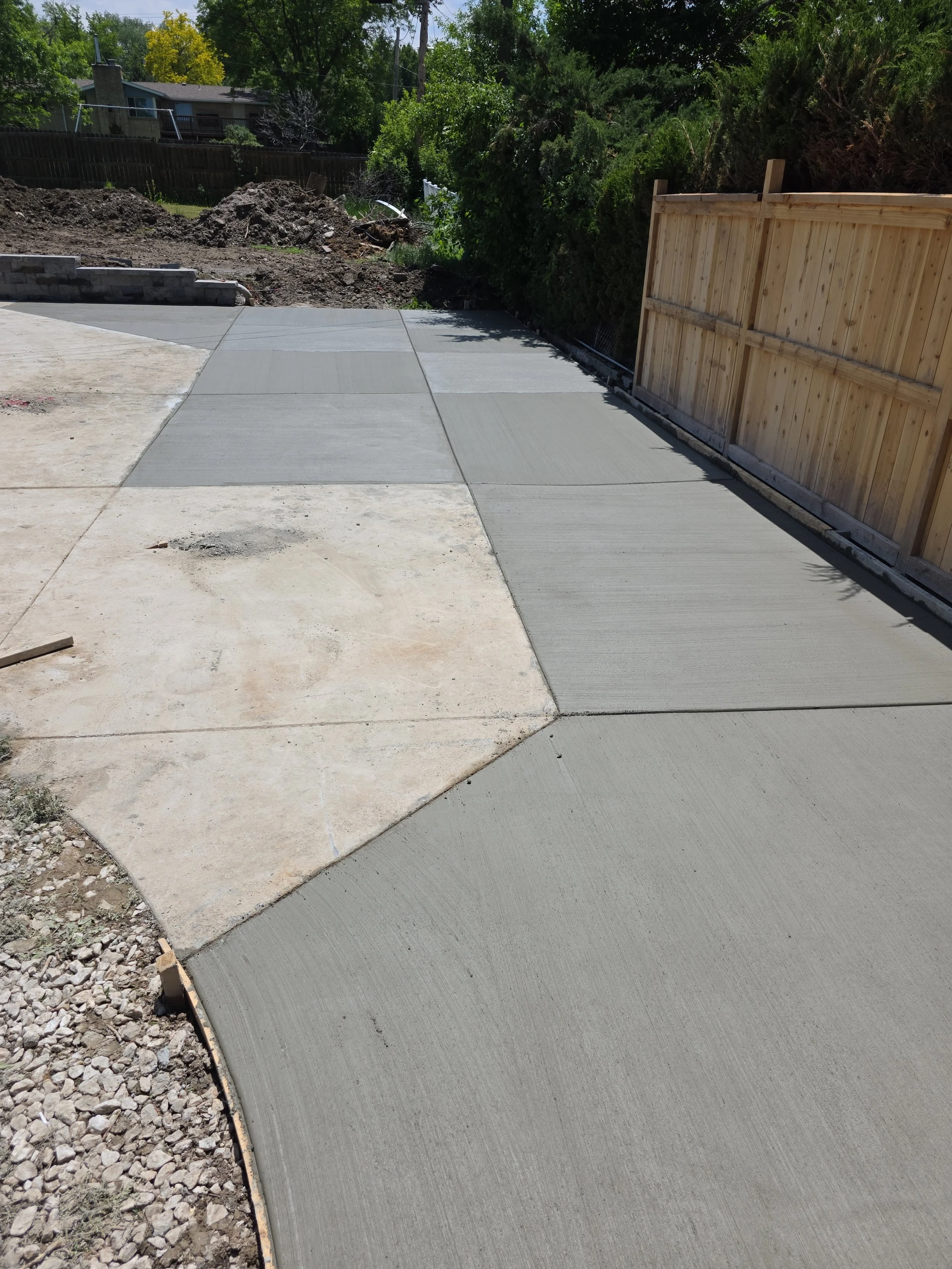 Newly poured concrete sidewalk with a wooden fence on the right and a dirt yard on the left. Green trees and a house are visible in the background.