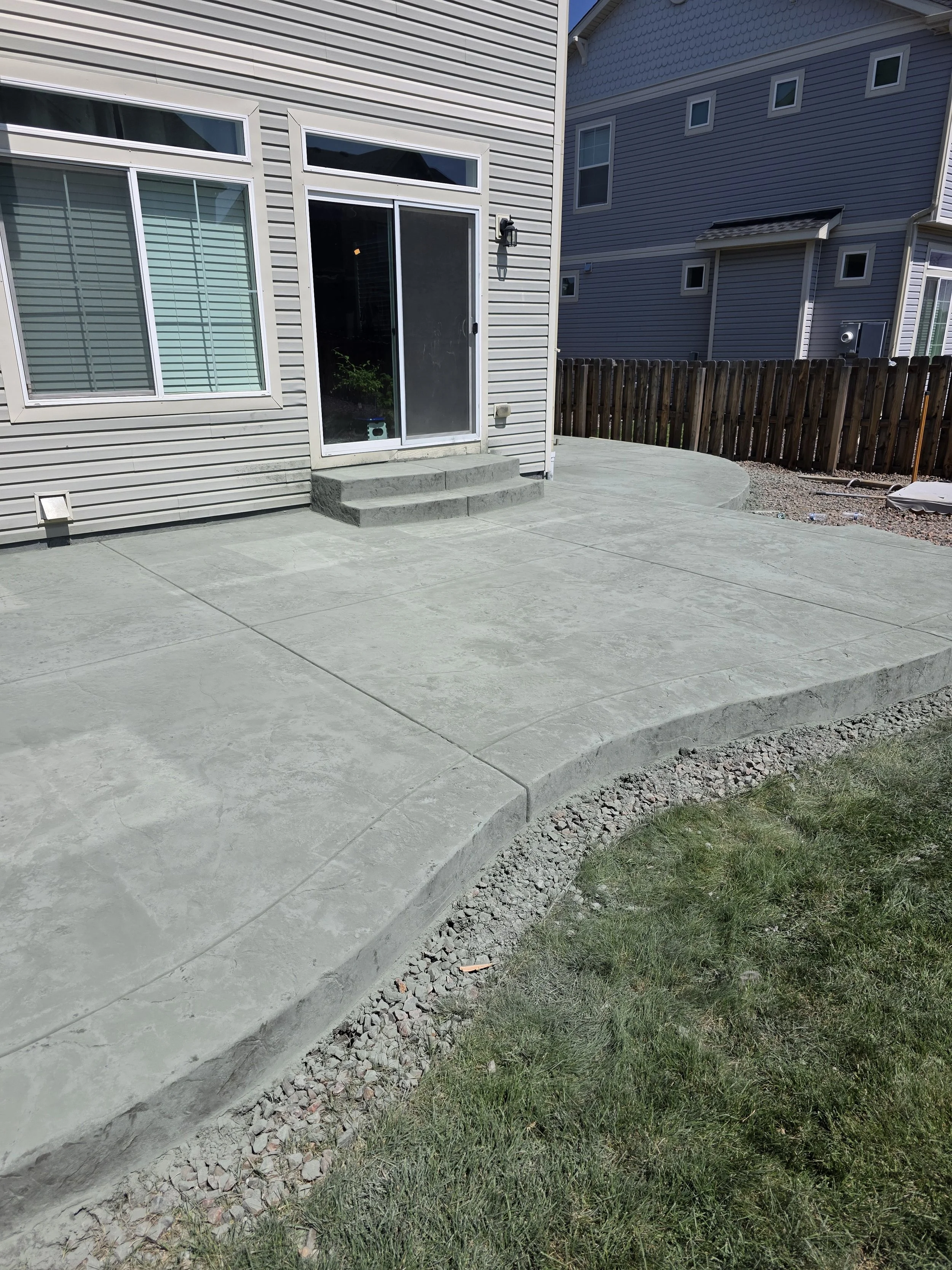 A backyard concrete patio with steps leading up to a sliding glass door, a wooden fence, and a small patch of grass on the right, with neighboring houses in the background.