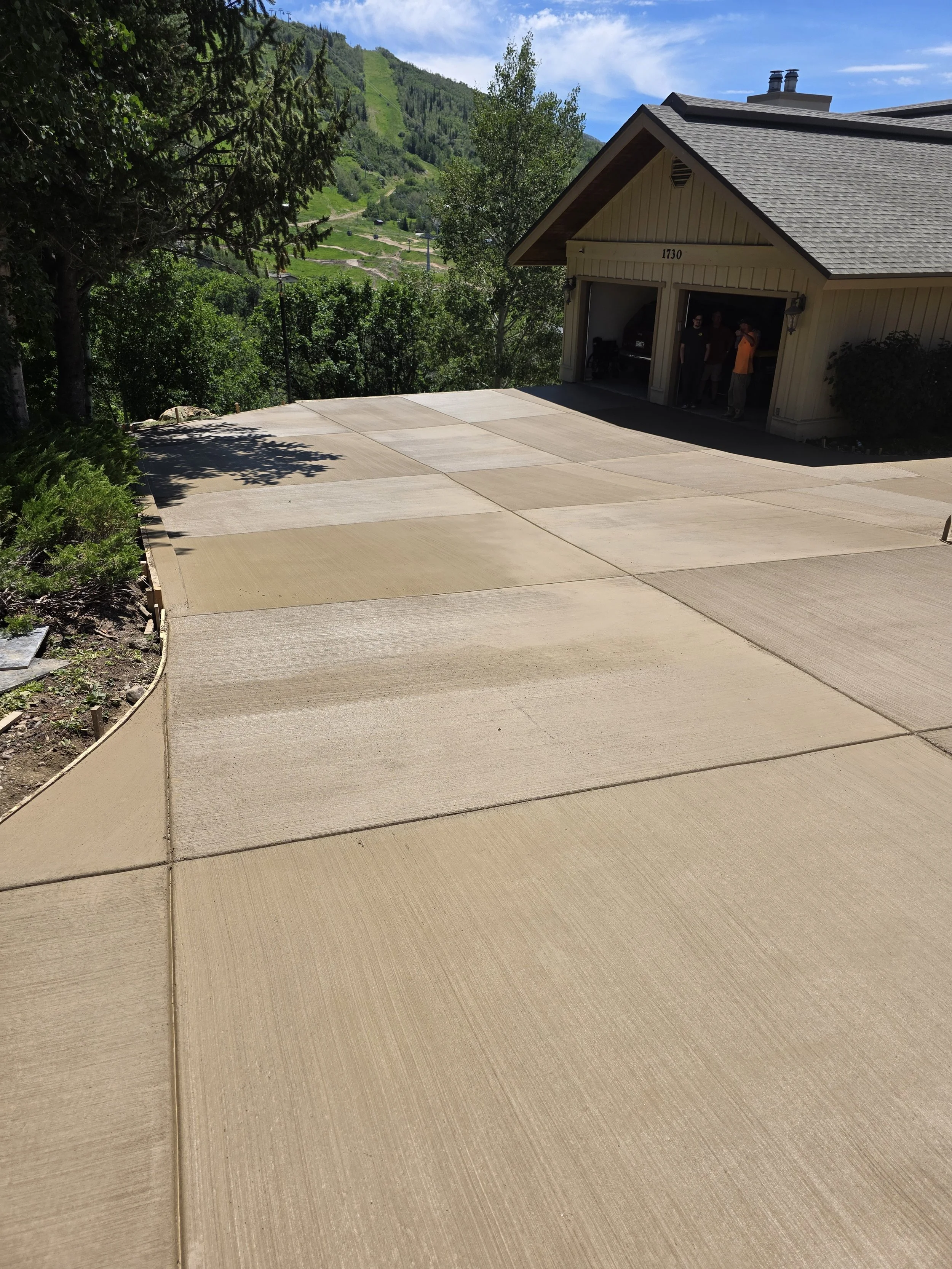 Newly paved concrete driveway leading to a beige garage with three people standing inside, surrounded by lush green trees and mountainous terrain in the background.