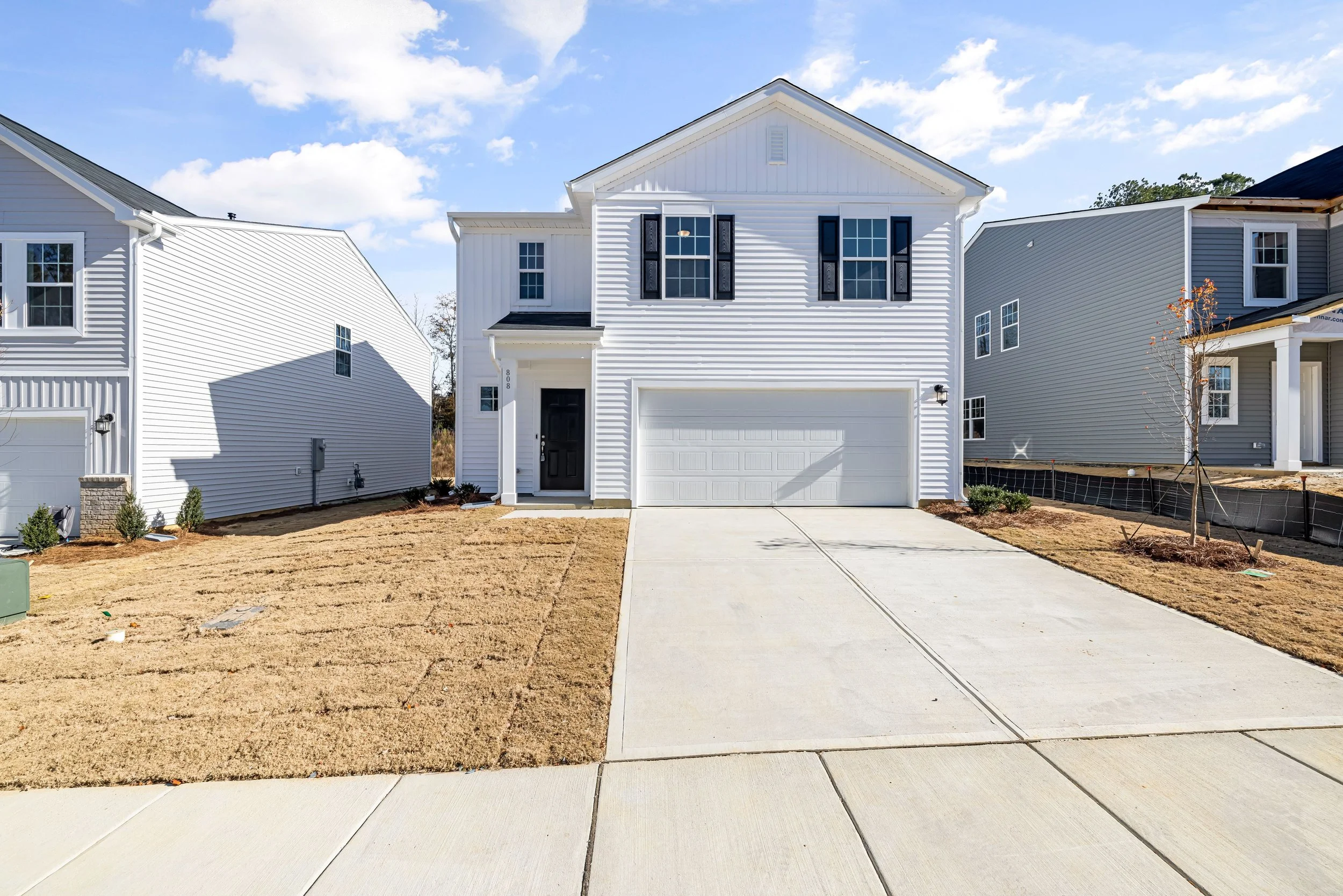 A white two-story house with black shutters and a black front door, surrounded by a partially finished lawn and a driveway under a blue sky with scattered clouds.