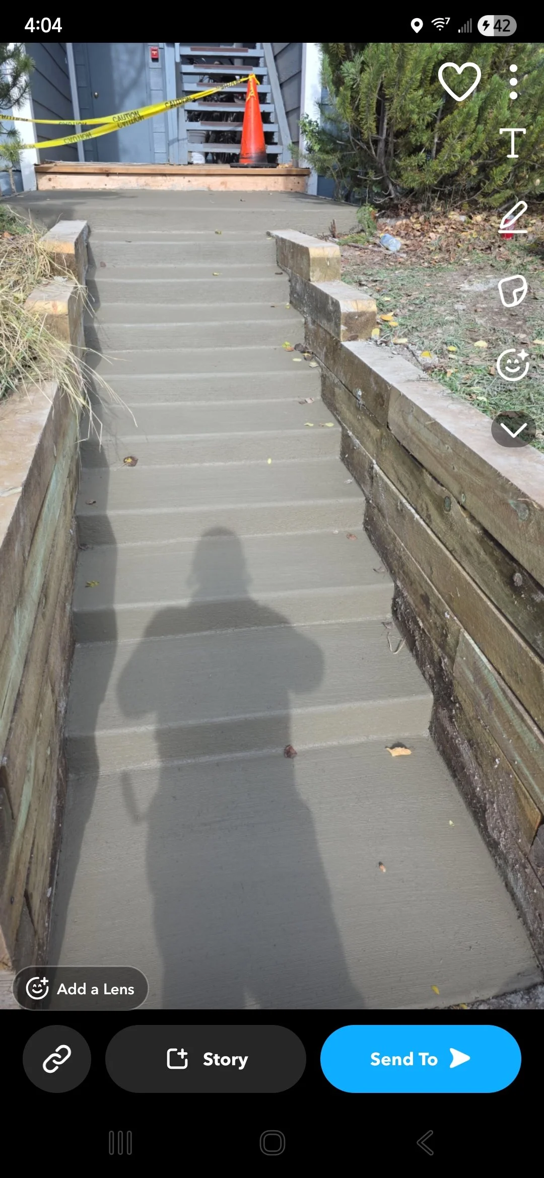 Newly poured concrete sidewalk with stairs leading up to a door, bordered by wooden planks and brick sides. Caution tape and an orange cone are at the top of the stairs. Vegetation and discarded plastic bottle are on the right.