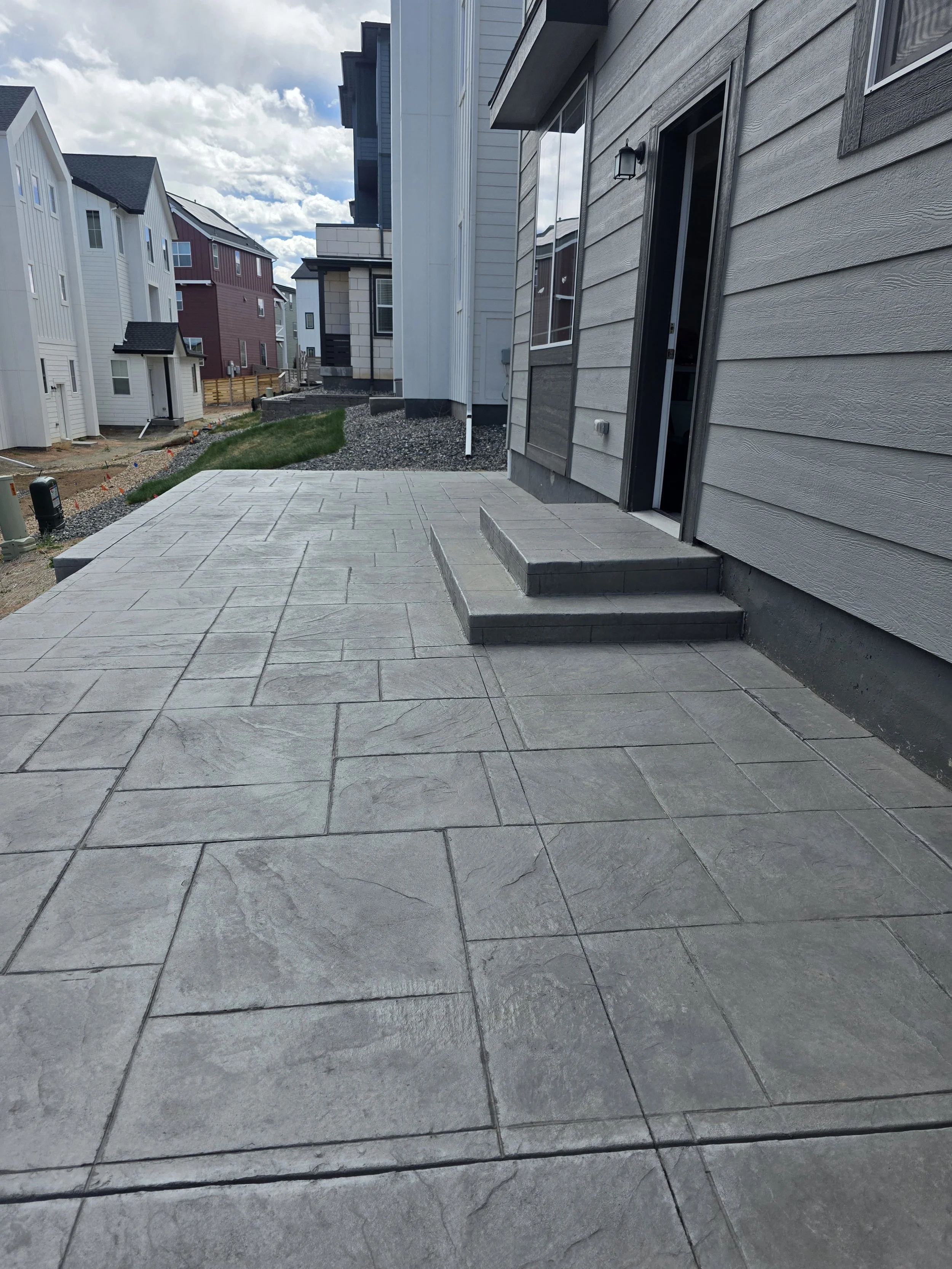 Rear view of new houses with various exterior siding, some with gray and white siding, and a small concrete patio in the foreground under a partly cloudy sky.