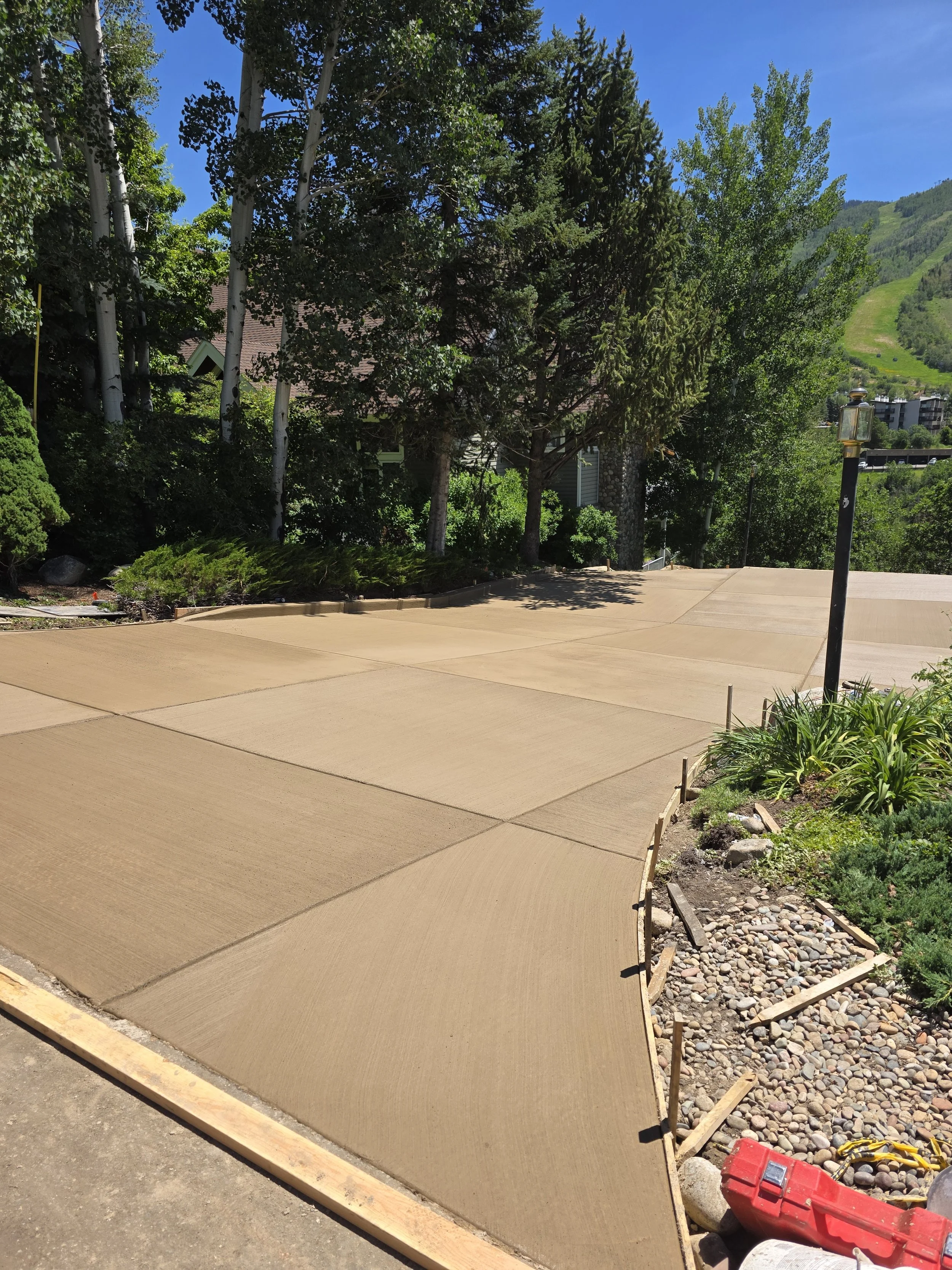Freshly poured concrete sidewalk with framing edges, garden bed with rocks and plants on right, trees and green mountains in background under blue sky.
