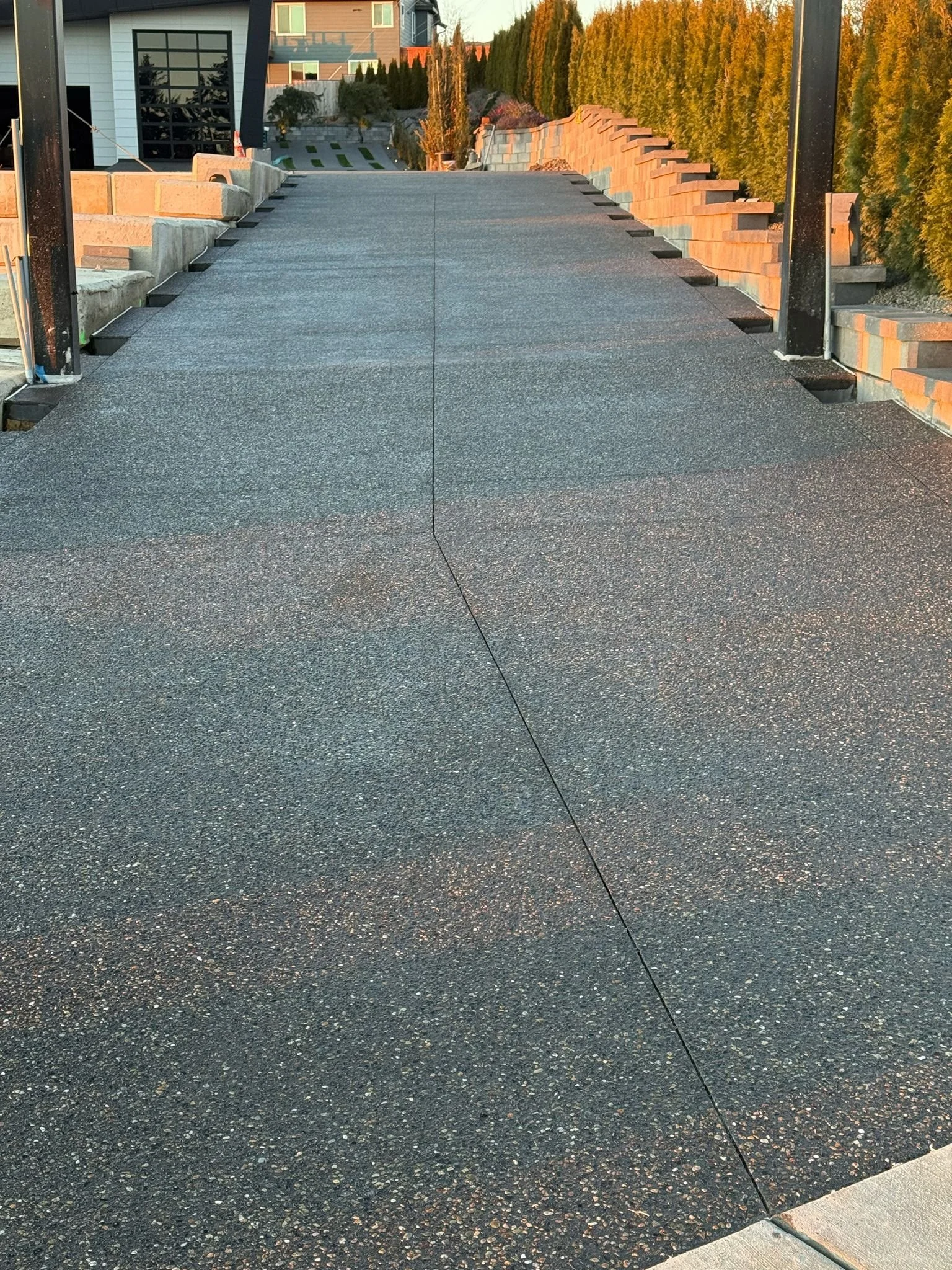 View of a black asphalt road or driveway with small multicolored stones, leading to a modern house and lined with trees and brick walls at sunset.