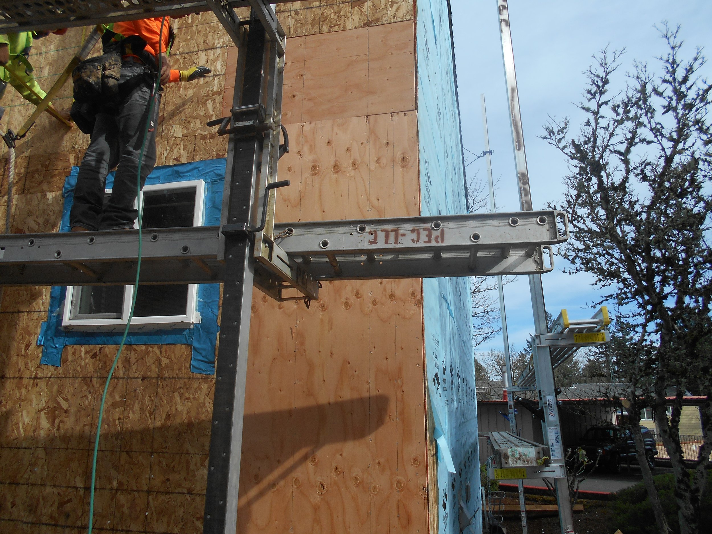 Construction workers on scaffolding working on the exterior wall of a building, with a partially installed window, in a residential neighborhood with trees and houses nearby.