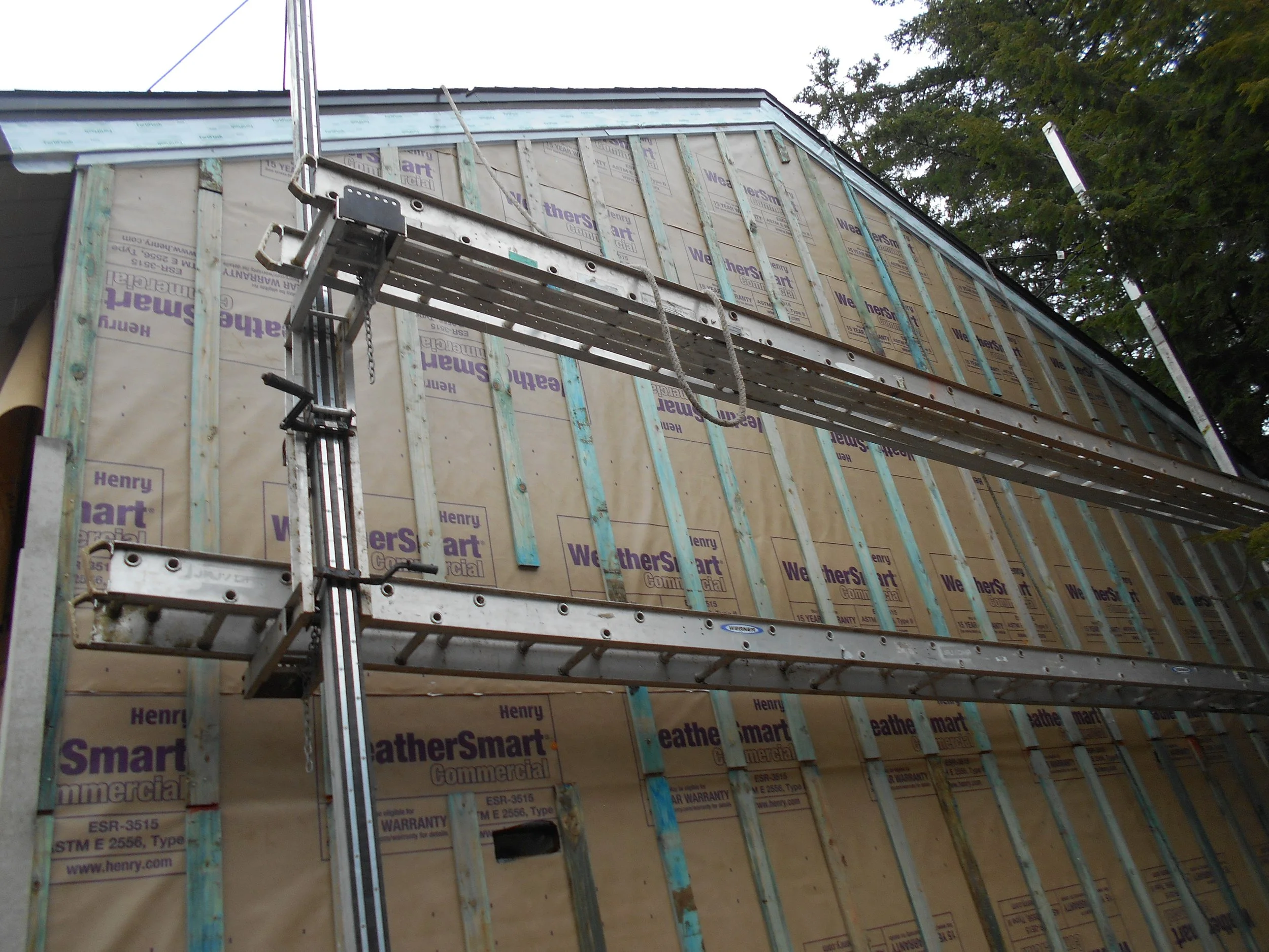 A house under construction, with visible sheathing and framing, a scaffold for workers, and a chimney on the roof surrounded by trees.