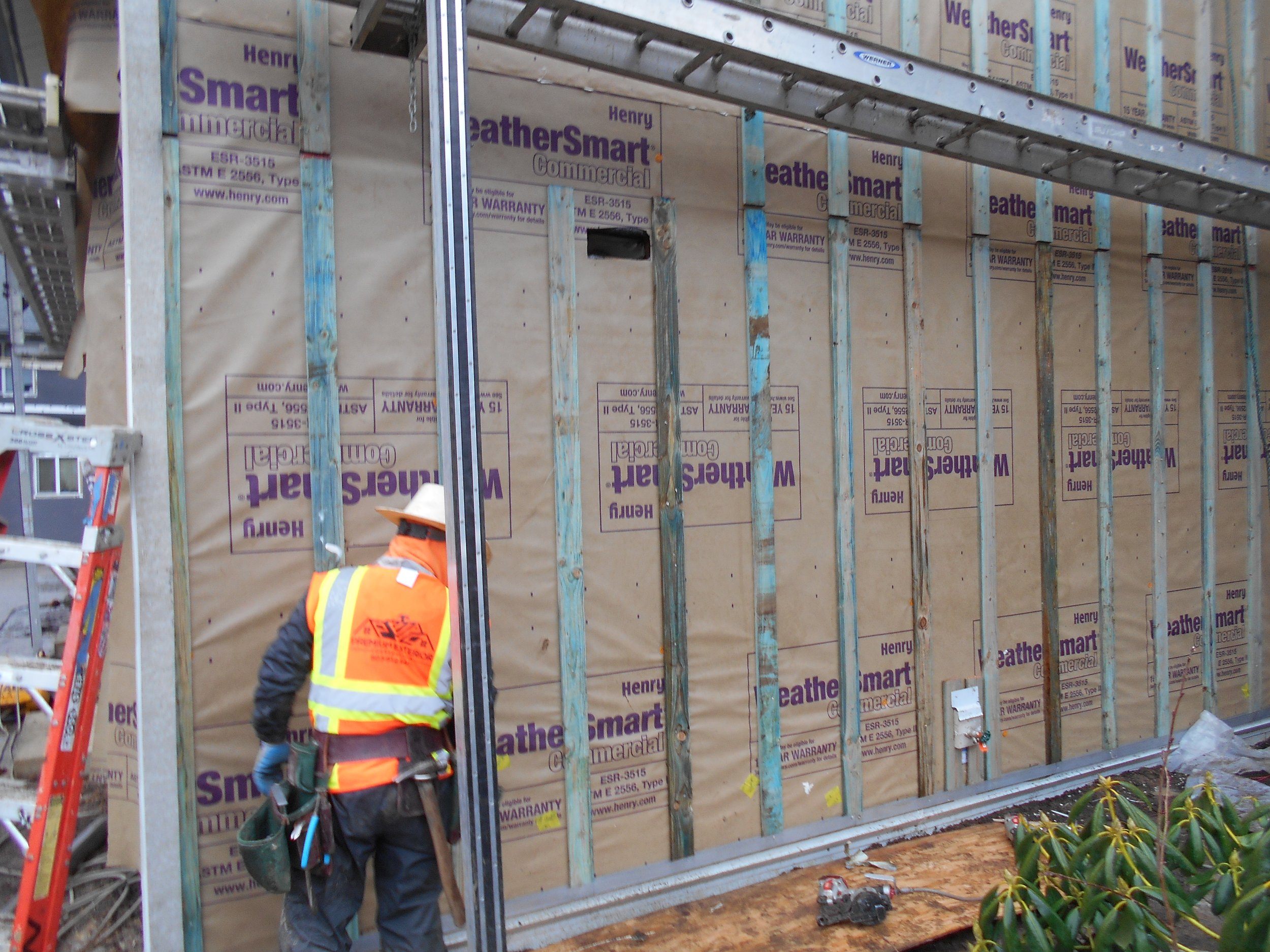 Construction worker in safety vest and helmet installing siding on a house with metal framework and insulation panels.