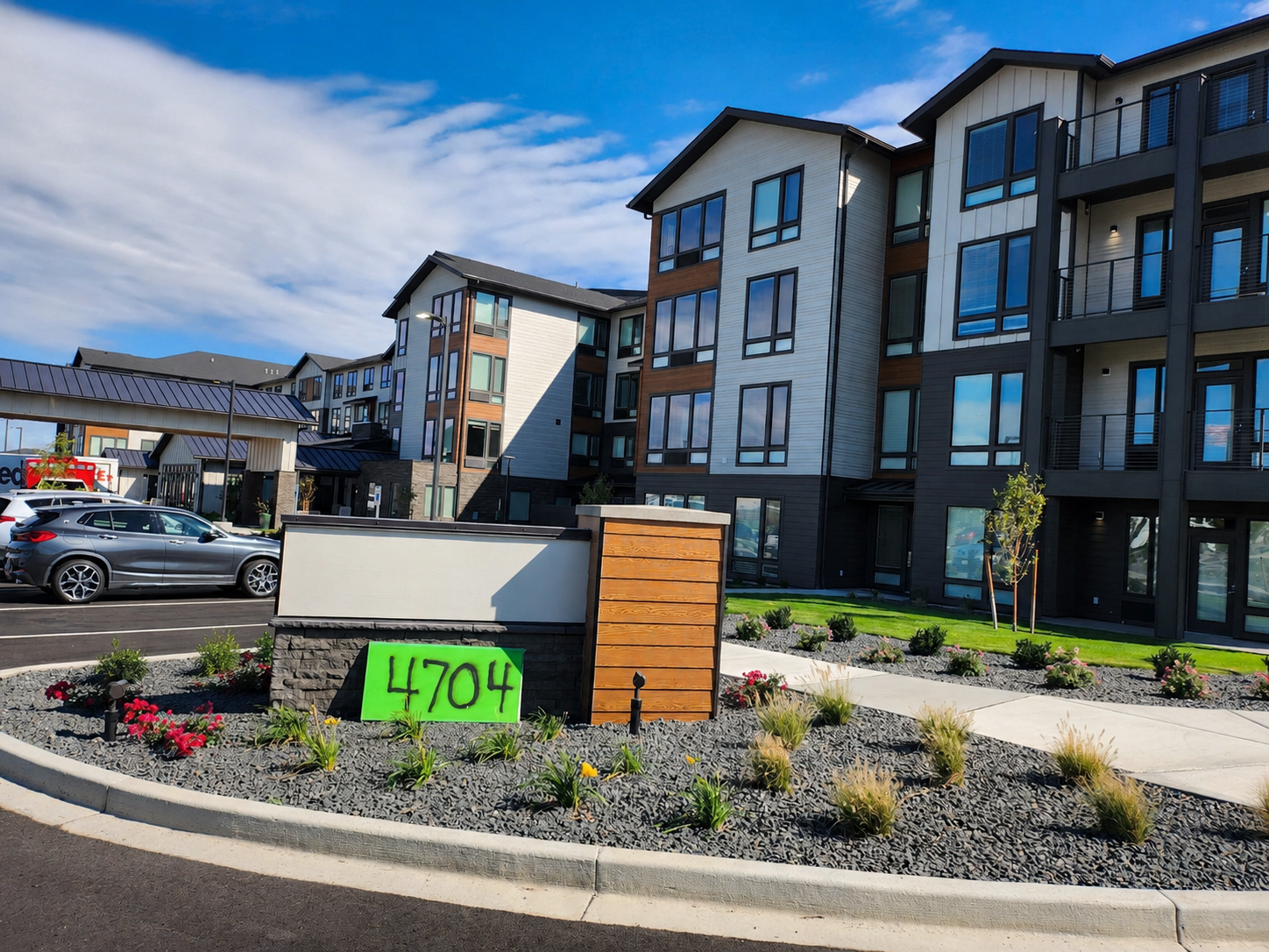 Modern apartment complex with multiple stories, large windows, and a small landscaped area with flowers and shrubs in the foreground. A bright green sign with the number 4704 is displayed near the landscaped area. Several cars are parked along the street in front of the building.