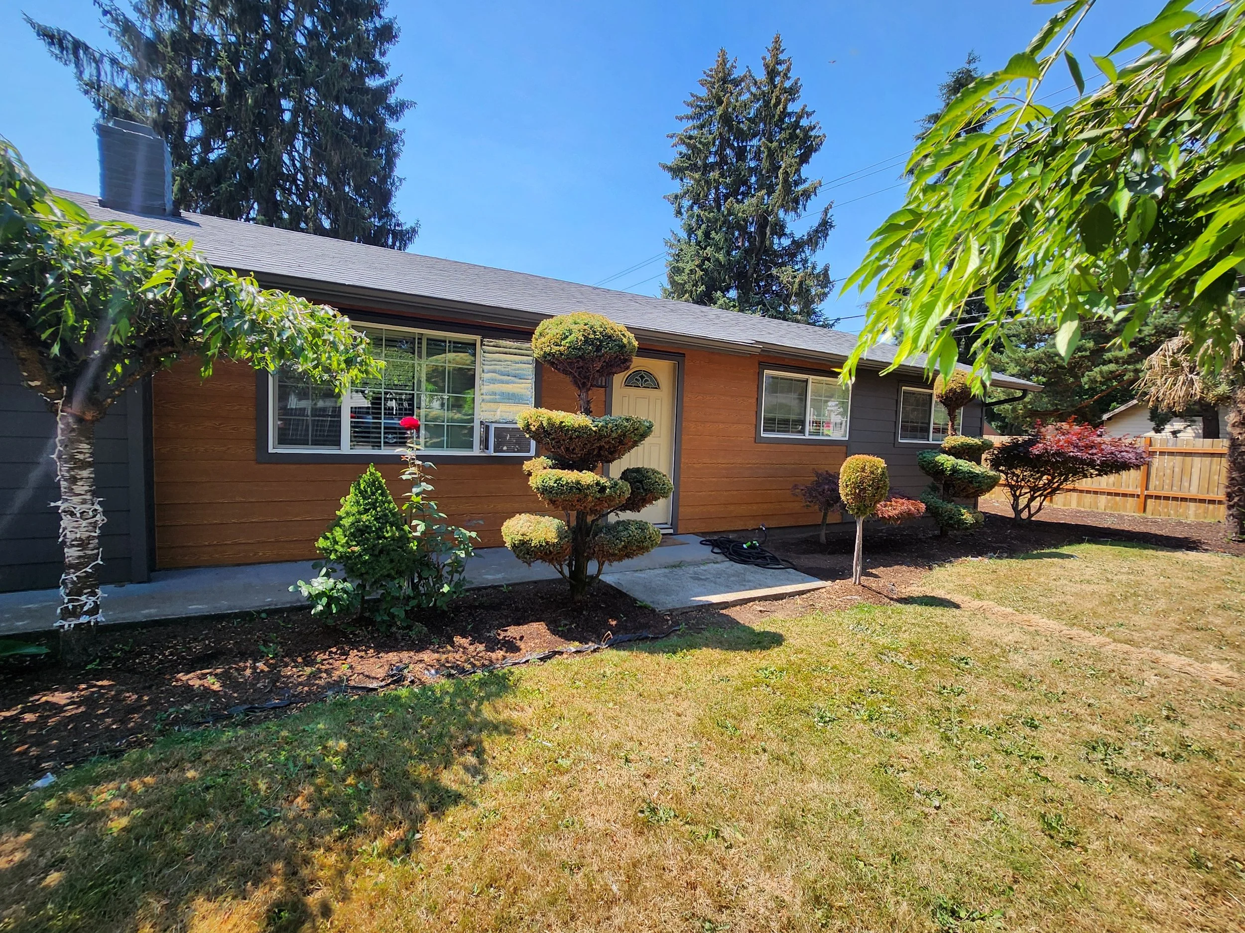 A house with brown and gray exterior, large windows, and a front door, surrounded by various shaped and sized pruned trees and bushes, under a clear blue sky.