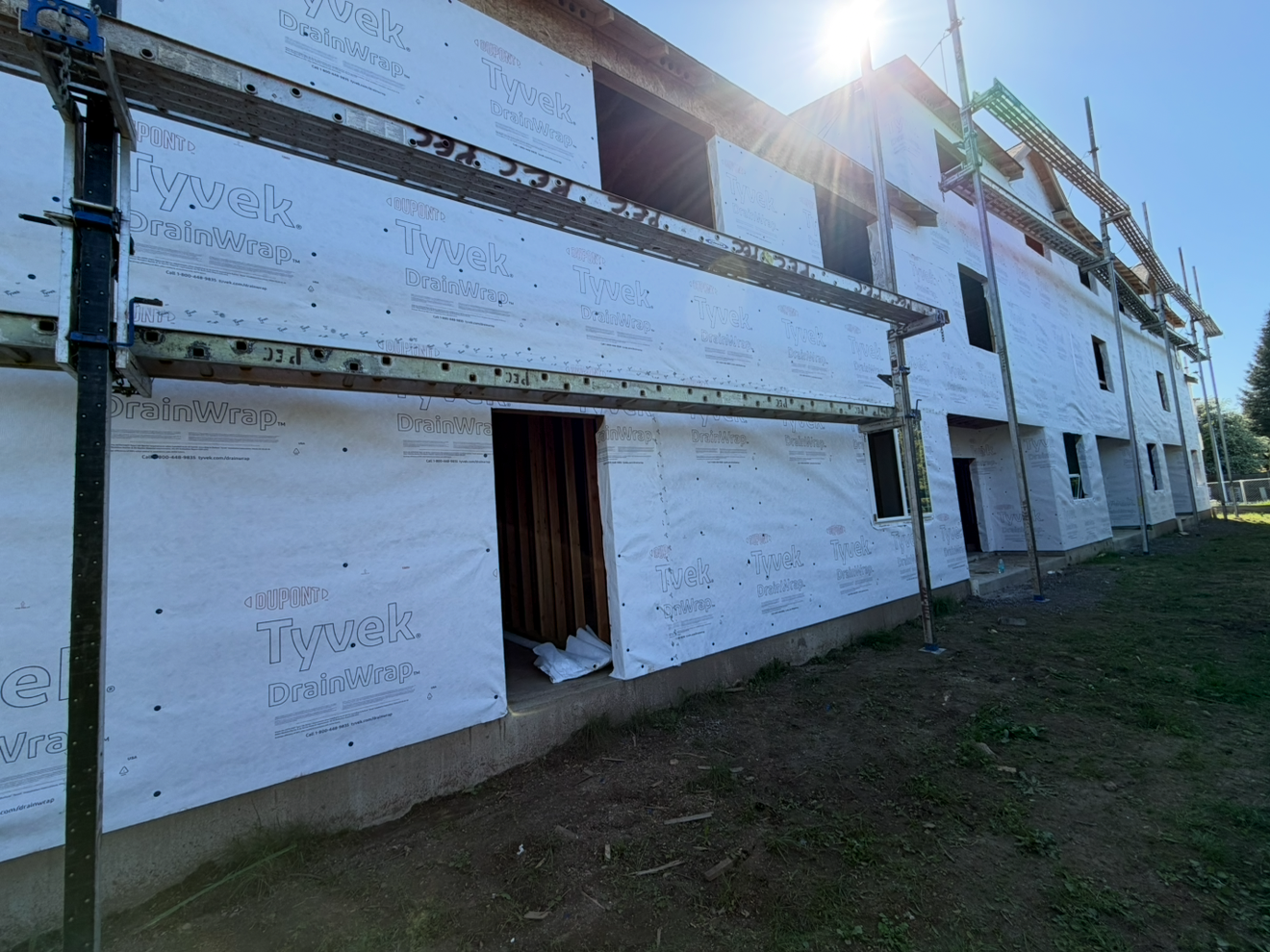 An under-construction multi-story building wrapped in white Tyvek DrainWrap with exposed window openings, scaffolding on the exterior, and a grassy ground in front, under a partly sunny sky.