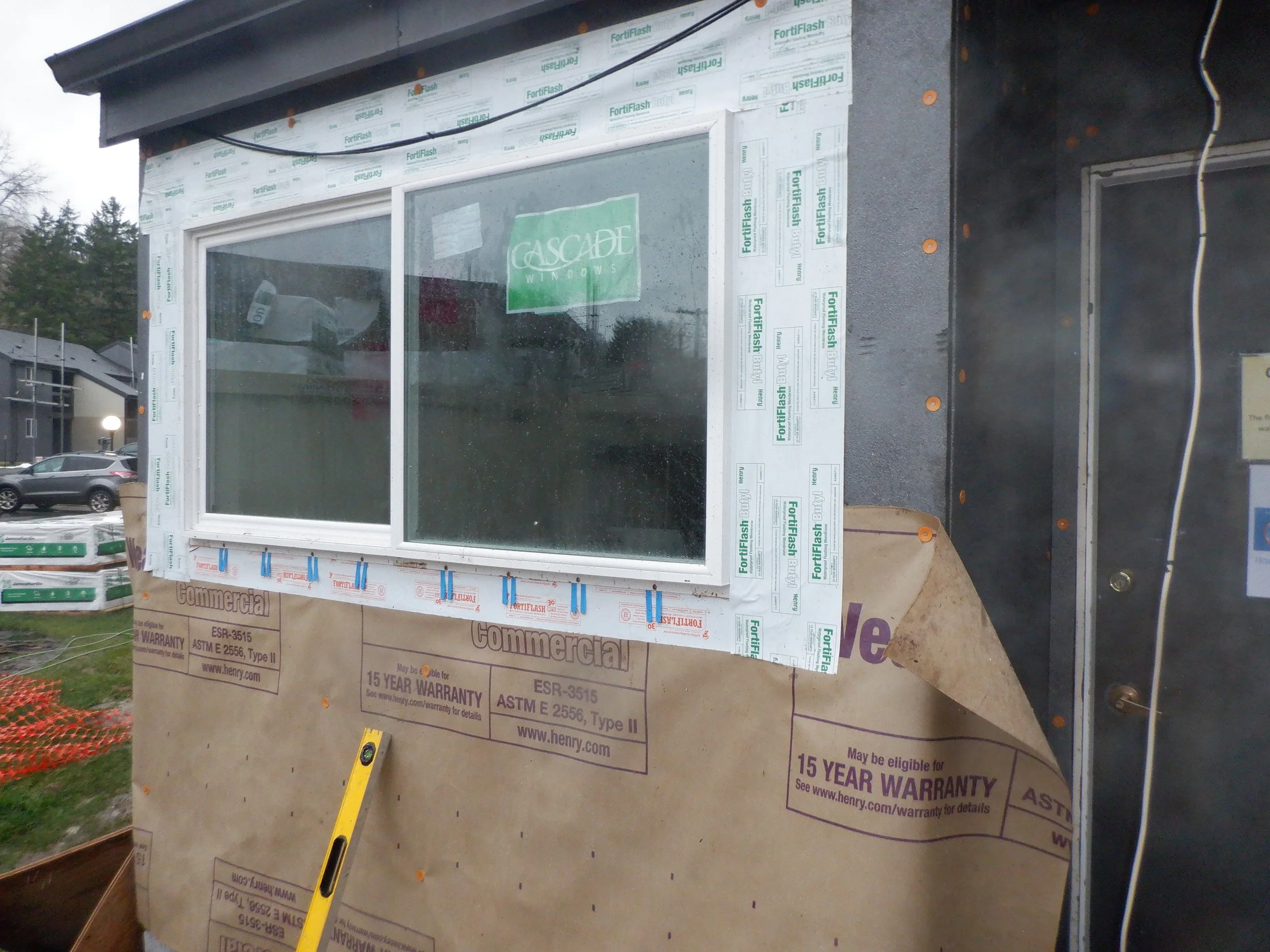 Construction site with a window frame installed, surrounded by weather-resistant sheathing with branding from Henry and FortiFlash, and a yellow level tool leaning against the wall.