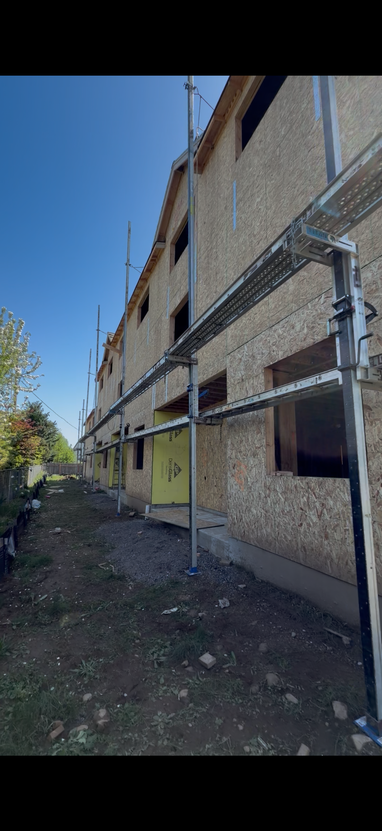 Building under construction with scaffolding and plywood sheathing, with a dirt yard and trees in the background.