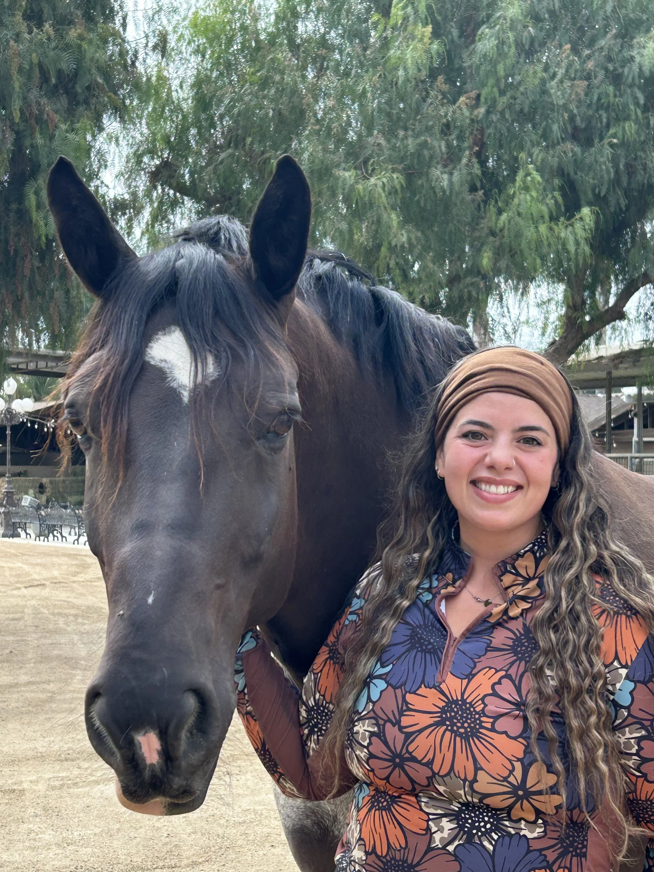 A woman with long wavy hair, wearing a colorful floral top and a brown headscarf, smiling beside a dark horse with a white star on its forehead in an outdoor setting with trees and a fence in the background.