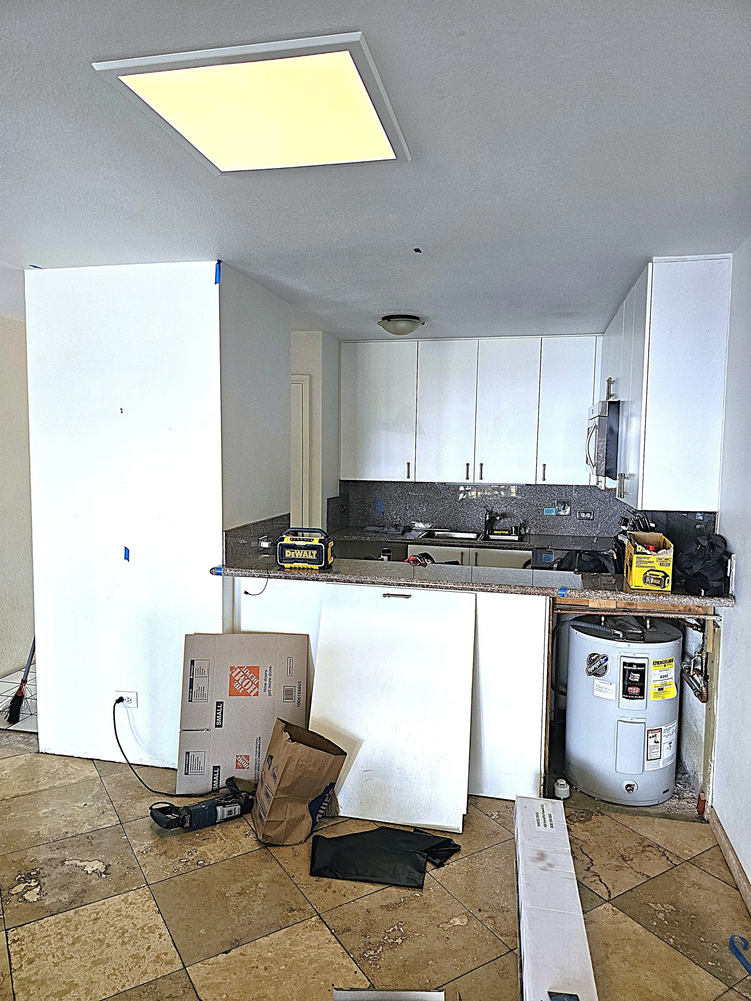 A kitchen under renovation with white cabinets, a granite countertop, a stainless steel sink, and a water heater. The floor with tiles, and there are various tools, boxes, and construction and demo materials.