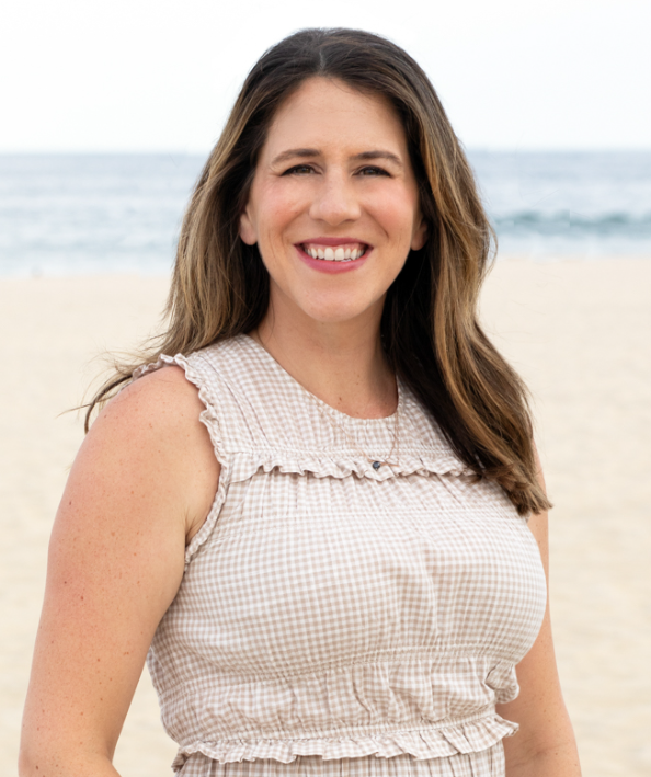 Woman with brown hair smiling at the beach, ocean in background.
