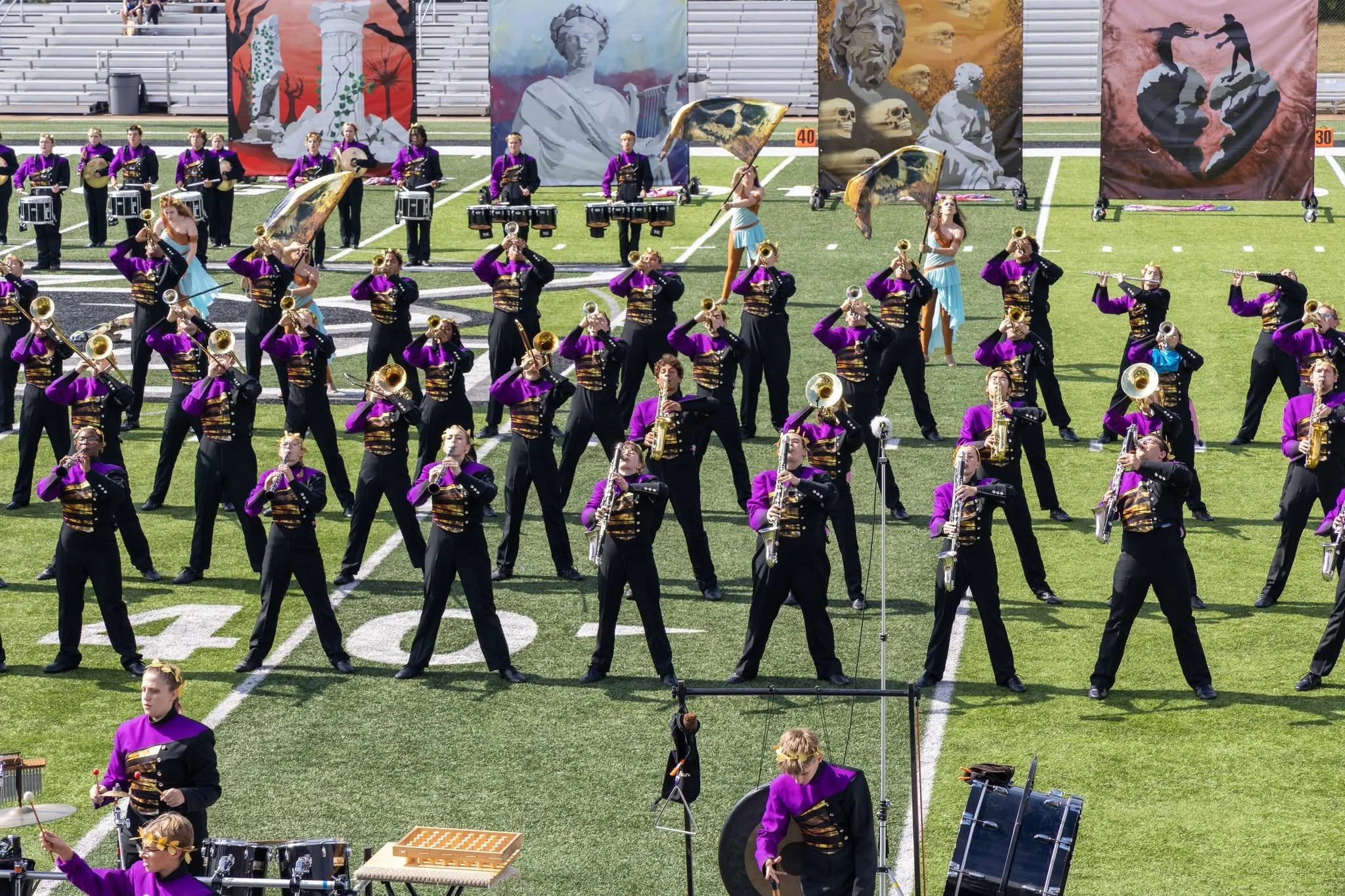 A marching band performing on a football field with large artistic banners in the background. The band members are wearing purple and black uniforms and are playing various musical instruments, including drums, saxophones, and trumpets.