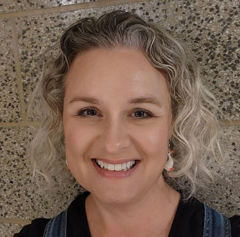 A woman with curly, blonde hair smiling at the camera, standing in front of a textured brick wall.