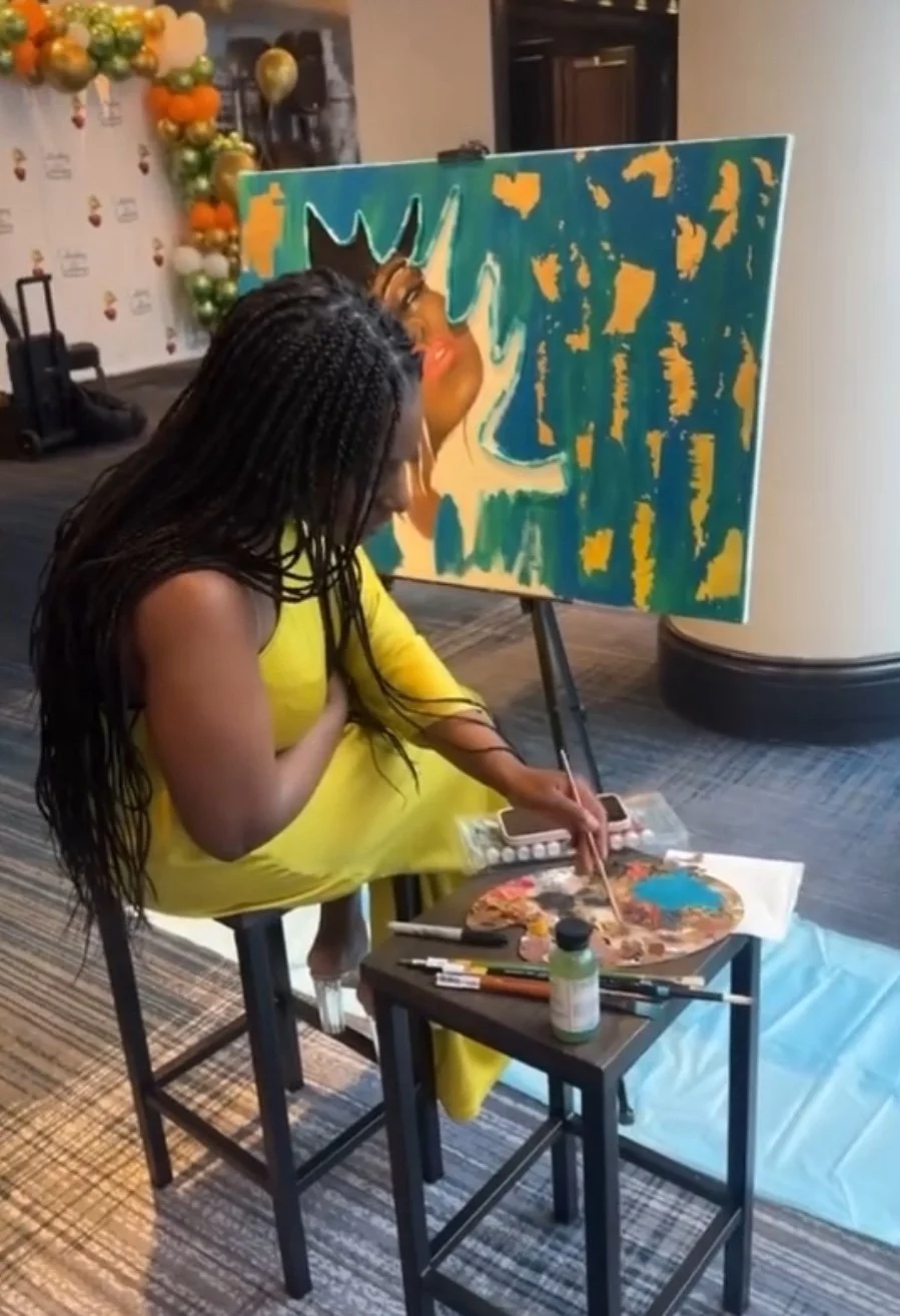 A woman in a yellow dress sits on a chair, painting an abstract portrait on a canvas with bright colors, while another person with long textured hair leans in to observe the artwork. Art supplies and a paint palette are on a small table next to them. In the background, there is a decorative balloon arrangement and a wall with festive branding.