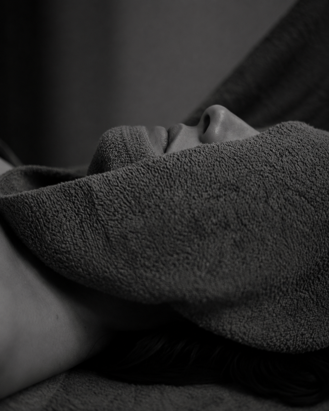 Close-up black and white photo of a person's face, partially covered by a textured fabric, with visible nose and lips, lying on a surface.