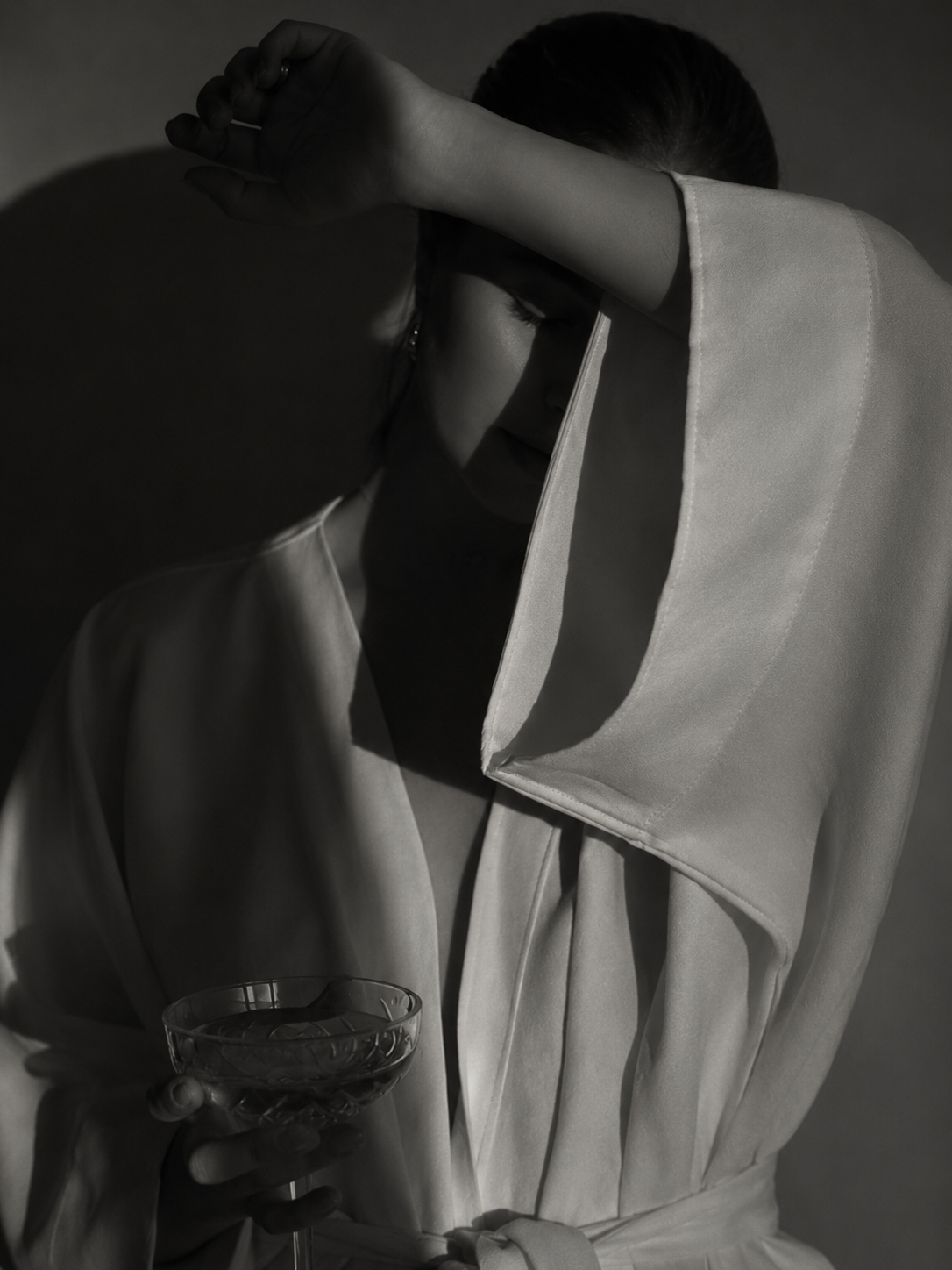 A woman in a satin robe with her eyes closed, holding a glass of water, shielding her face with her arm in a dramatic black and white portrait.