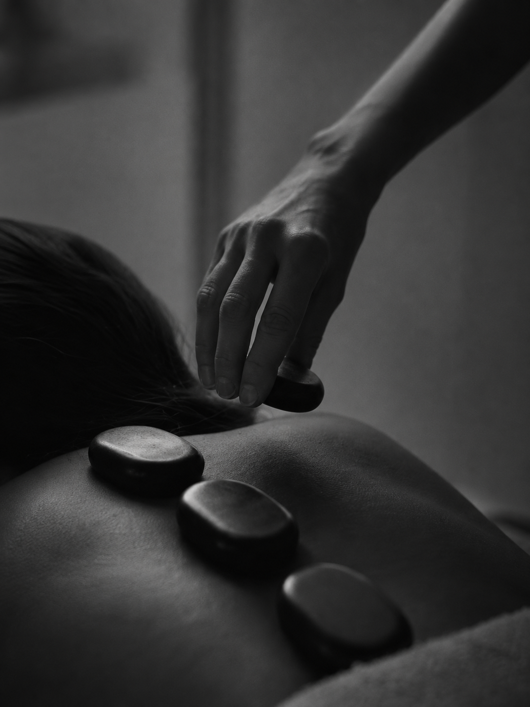 Black and white photo of a person receiving a hot stone massage, with smooth stones placed along their back, and a massage therapist’s hand gently pressing on one of the stones.