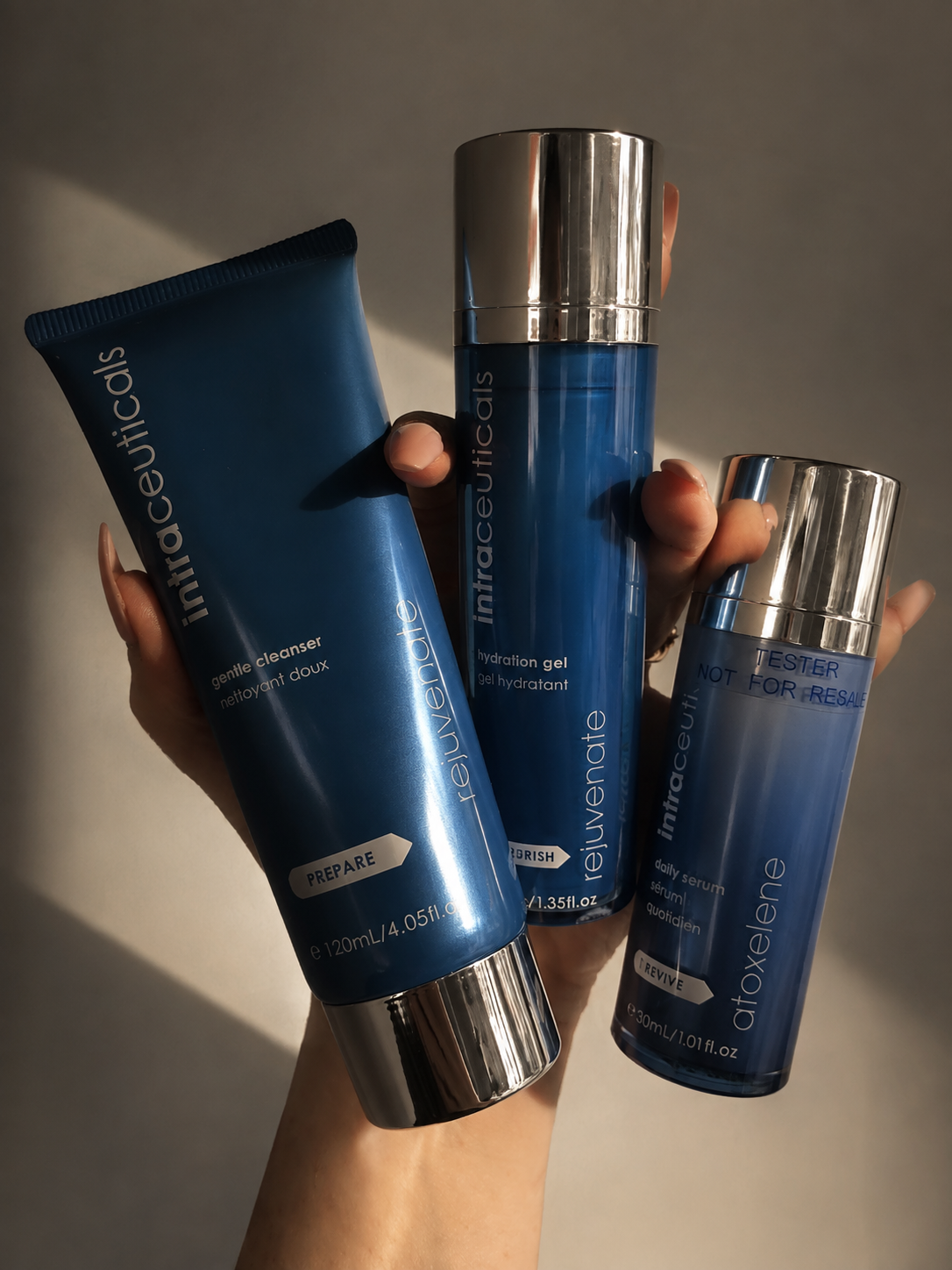 Hand holding three blue skincare bottles labeled Intraceuticals, including gentle cleanser, hydration gel, and daily serum. All have silver caps. The bottles are against a neutral background with shadow.