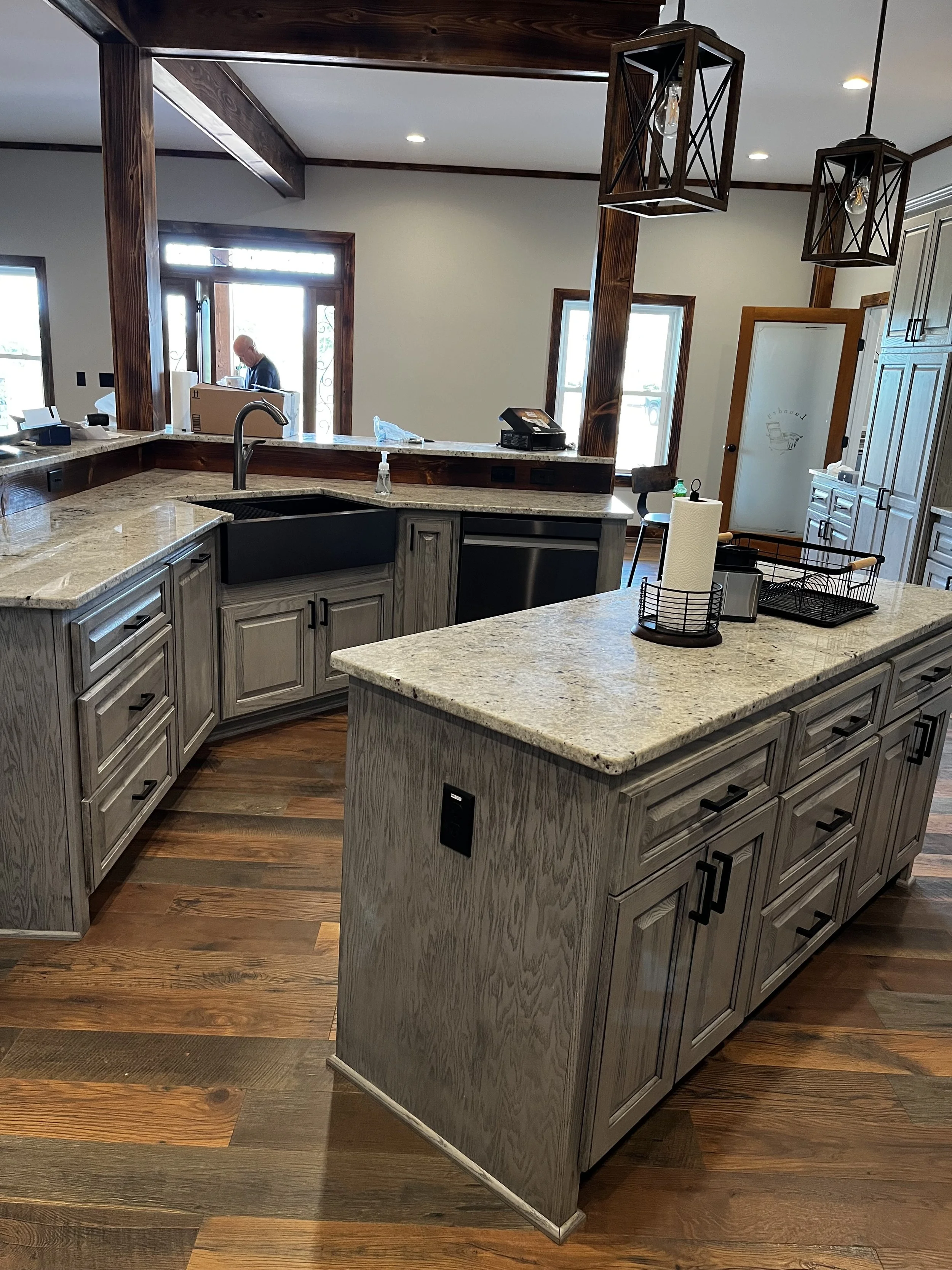 A modern kitchen with gray wooden cabinets, a large granite island with a built-in black sink, pendant lights, and a person with a bald head working near the front door.