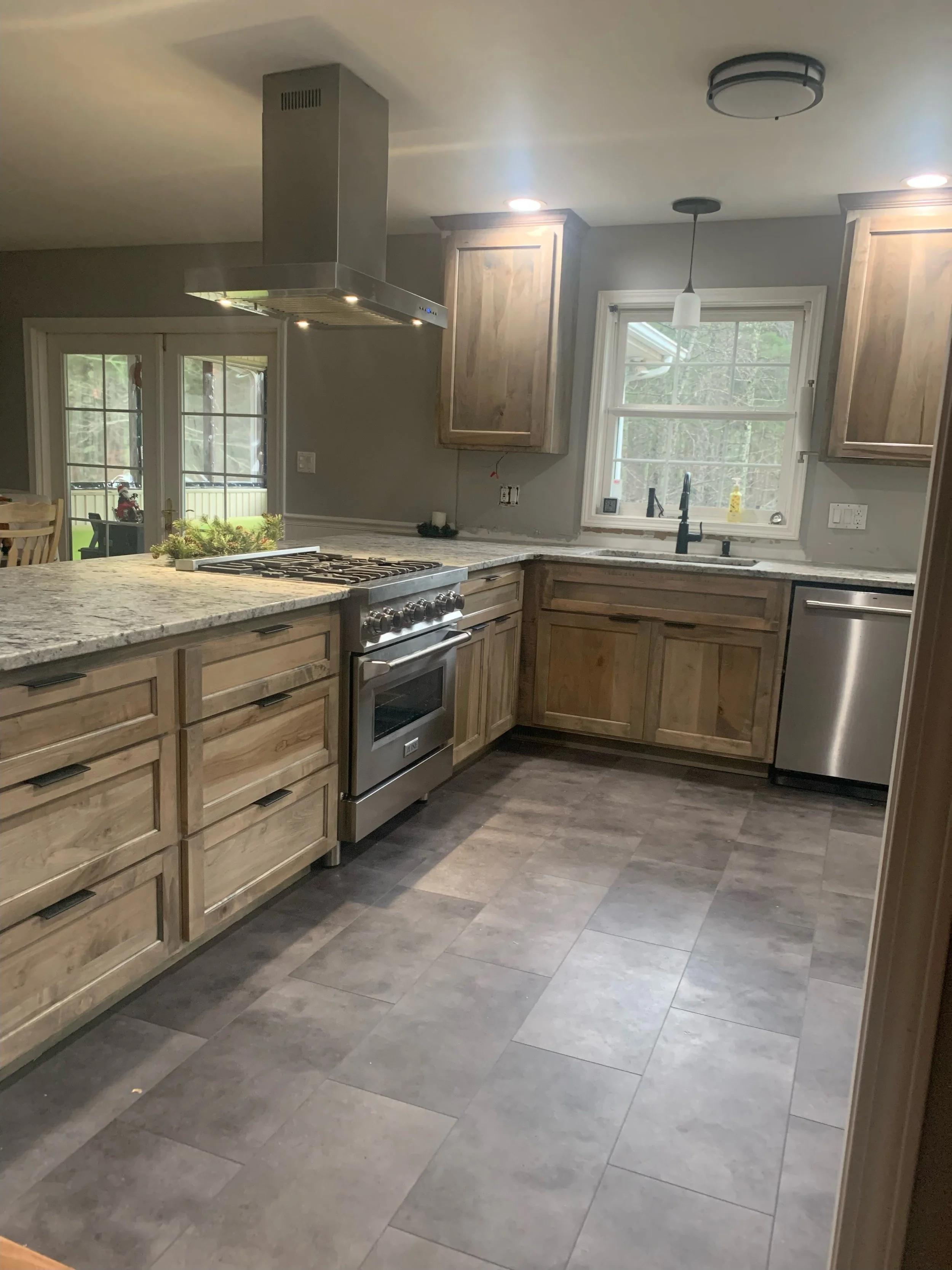 A kitchen with wooden cabinets, granite countertops, a stainless steel oven, and a sink under a window showing trees outside. There is a ceiling light fixture and a hanging pendant light.