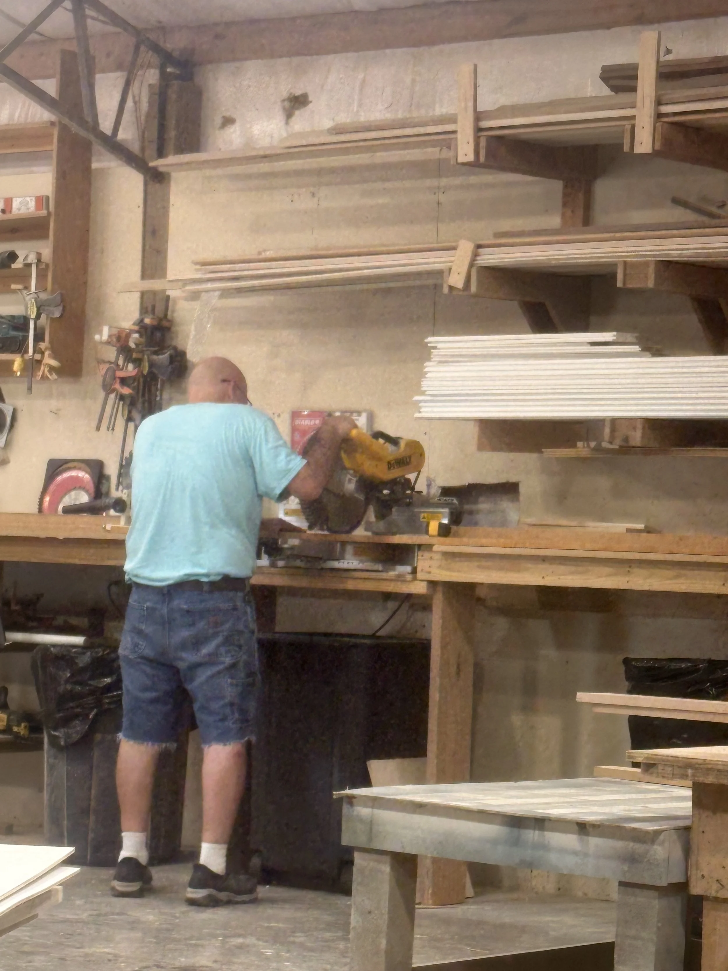 A man in a light blue shirt and shorts working with a yellow saw in a woodworking workshop. The workshop has wooden shelves, tools, and pieces of wood organized around.
