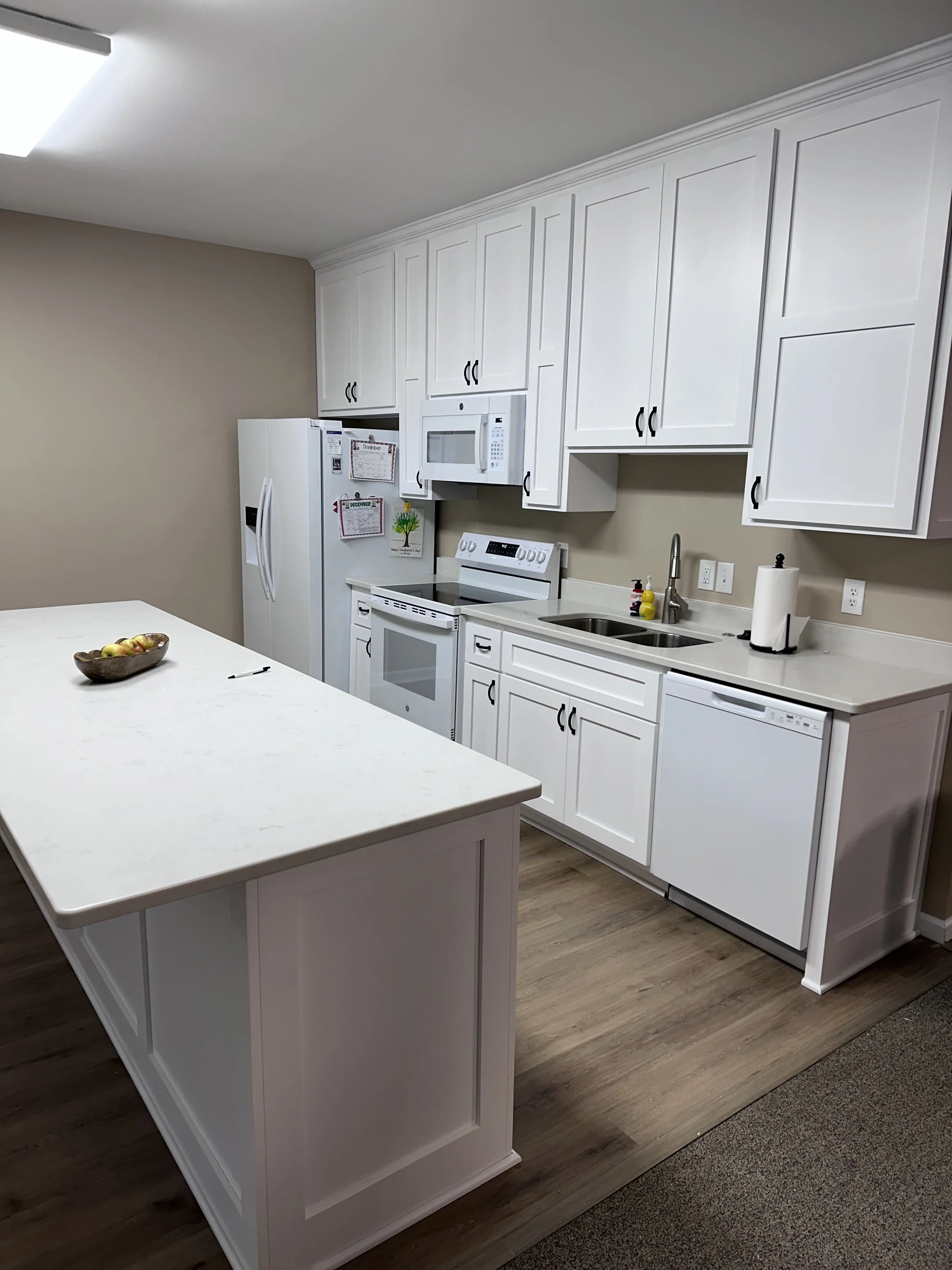 Modern kitchen with white cabinets, a white island, stainless steel sink, and a white refrigerator, microwave, and dishwasher, with dark wooden flooring.