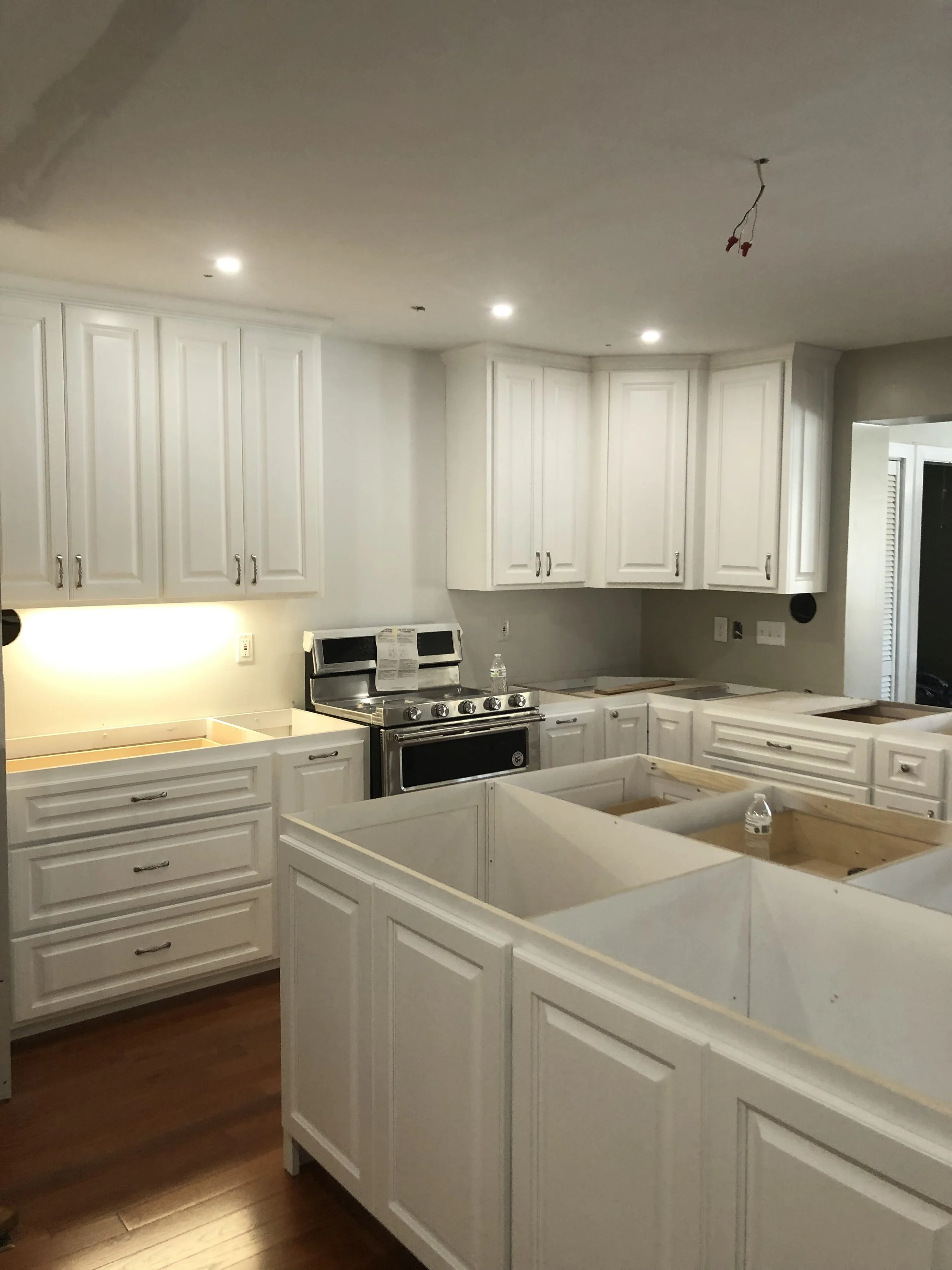 Kitchen under renovation with white cabinets, some removed drawers, and a stove, with construction materials and a water bottle on the counters.