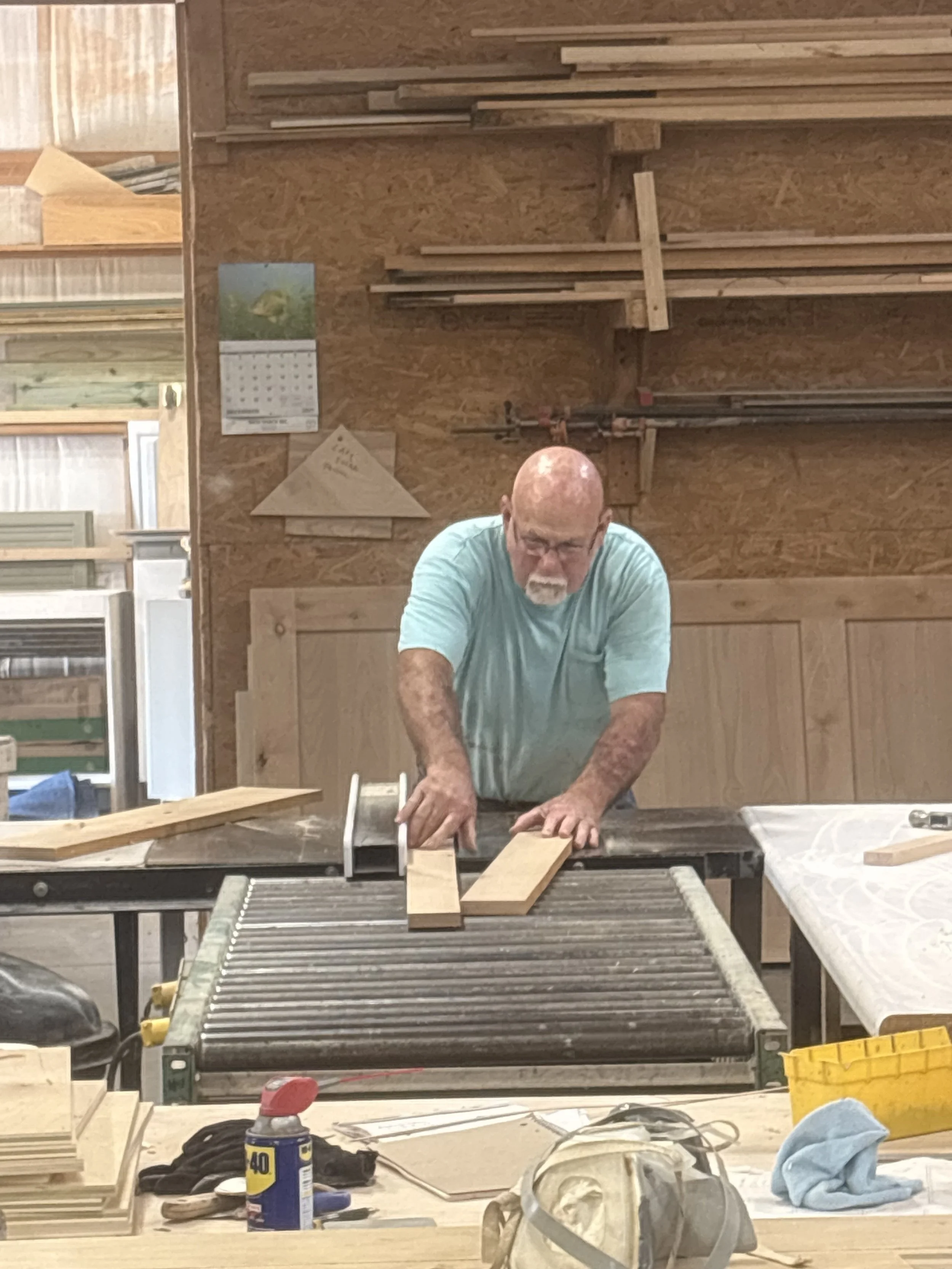 A man working in a woodworking shop, cutting a piece of wood on a table saw with various tools and materials scattered on the table, including a blue cloth, a yellow waste bin, and a can of glue.