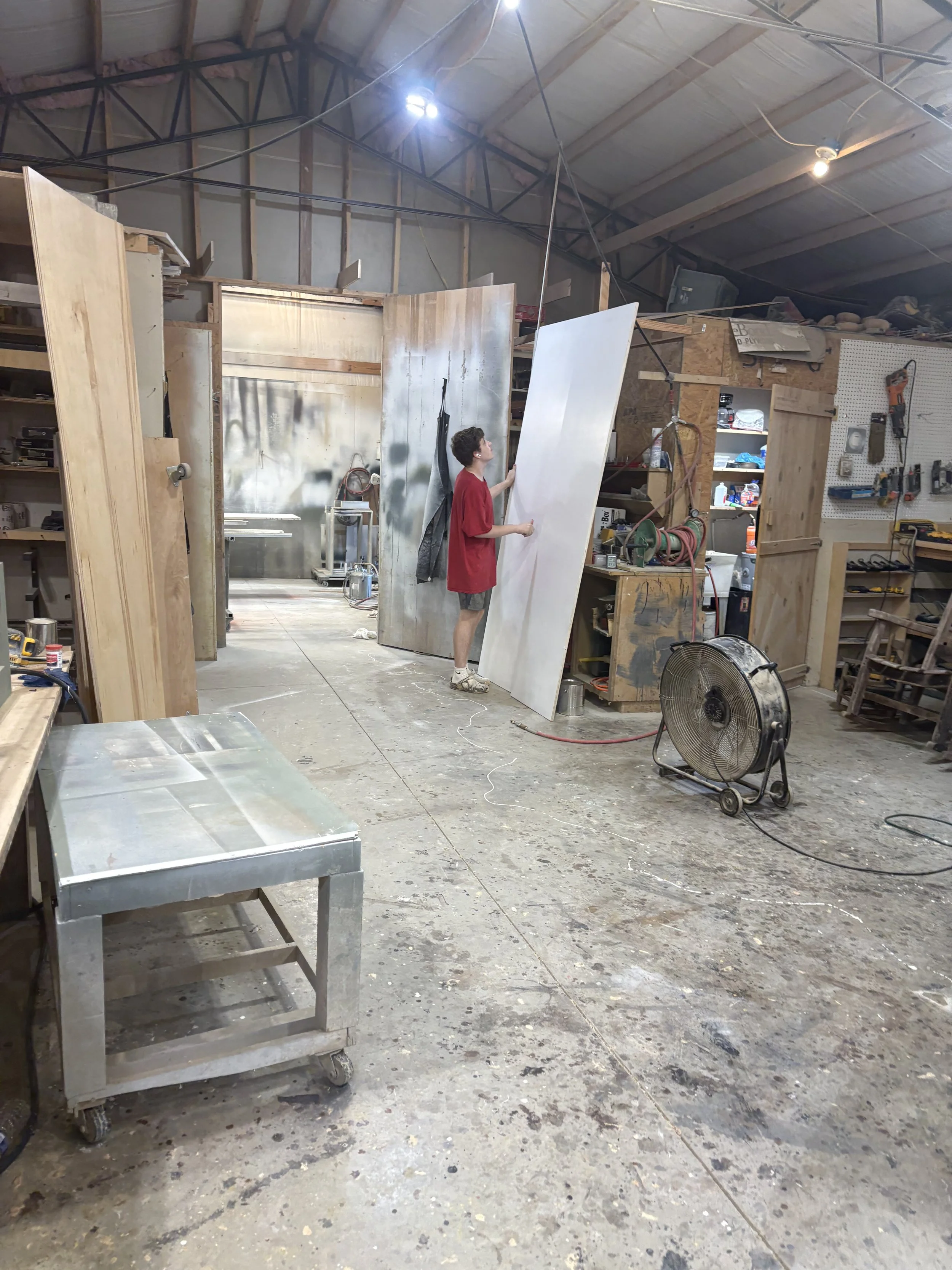 A person in a red T-shirt and shorts in a woodworking shop, holding a large piece of white material, possibly a panel or board. The shop is filled with various tools and equipment, with unfinished walls and a concrete floor.