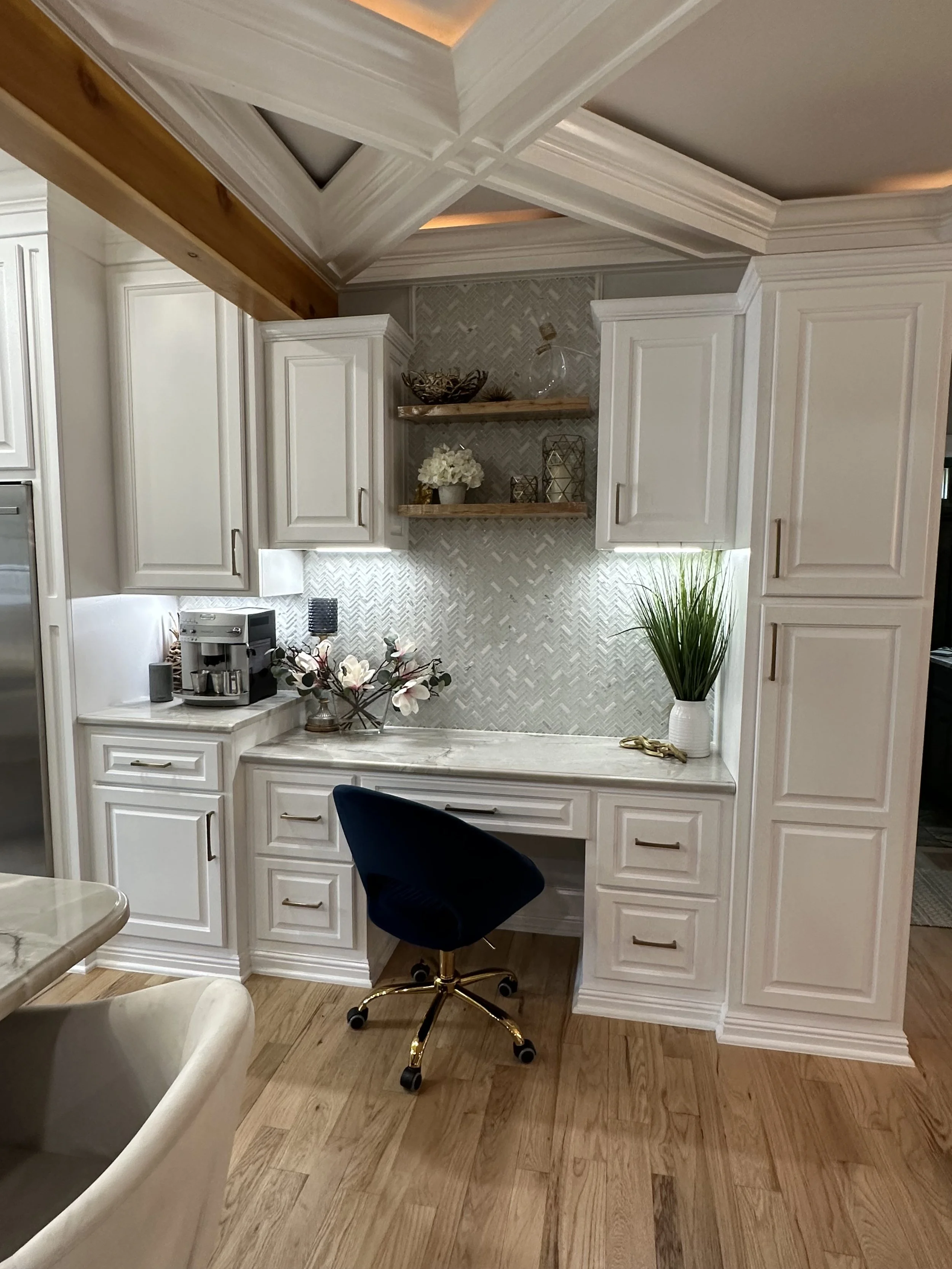 A kitchen with white cabinetry and a marble countertop, decorative shelves with plants, flowers, and glass objects, a black swivel chair with gold accents, and hardwood flooring.