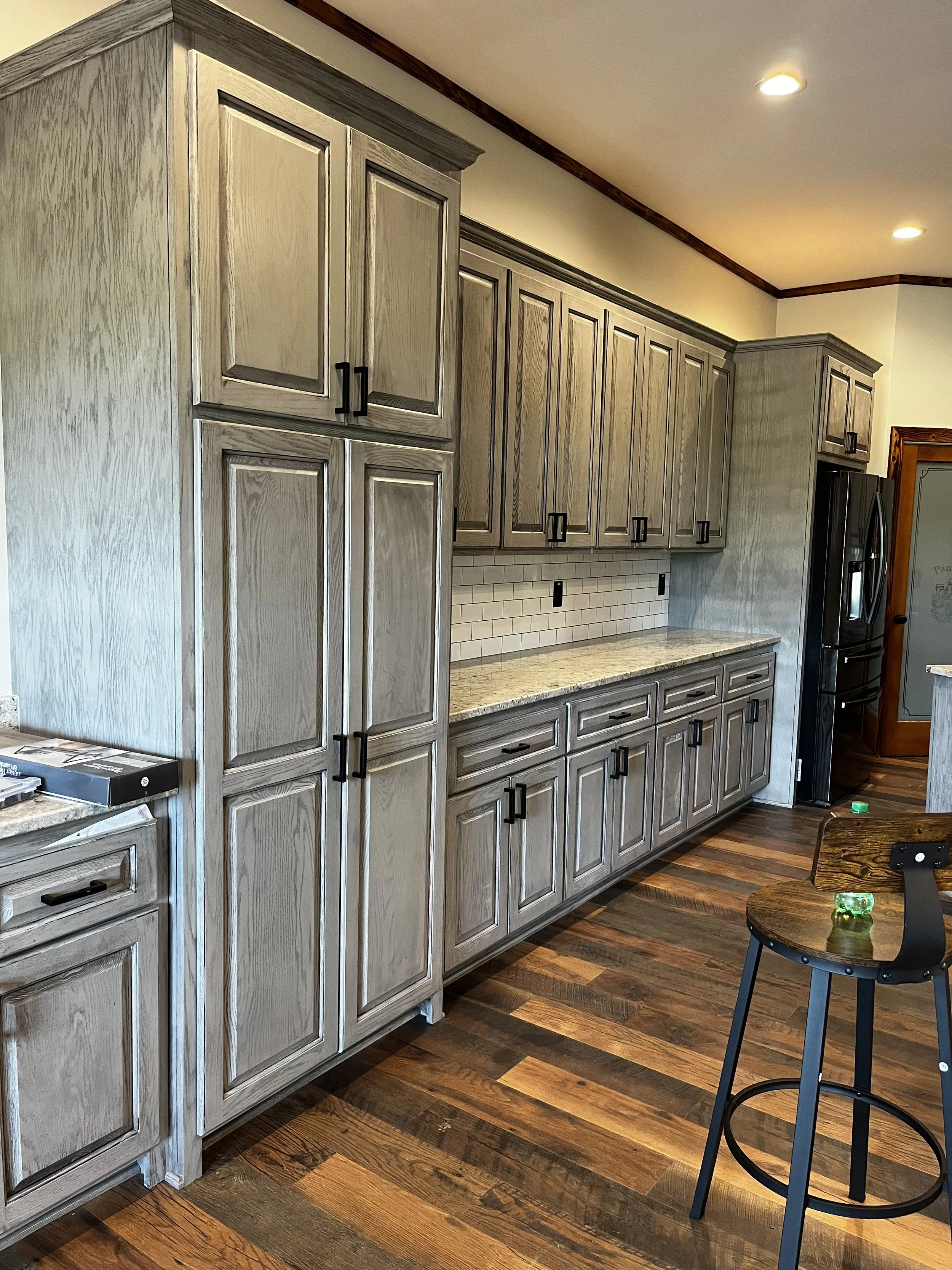 Kitchen with light wood cabinets, granite countertop, white tile backsplash, dark wood flooring, and a round bar stool with a green candle on it.