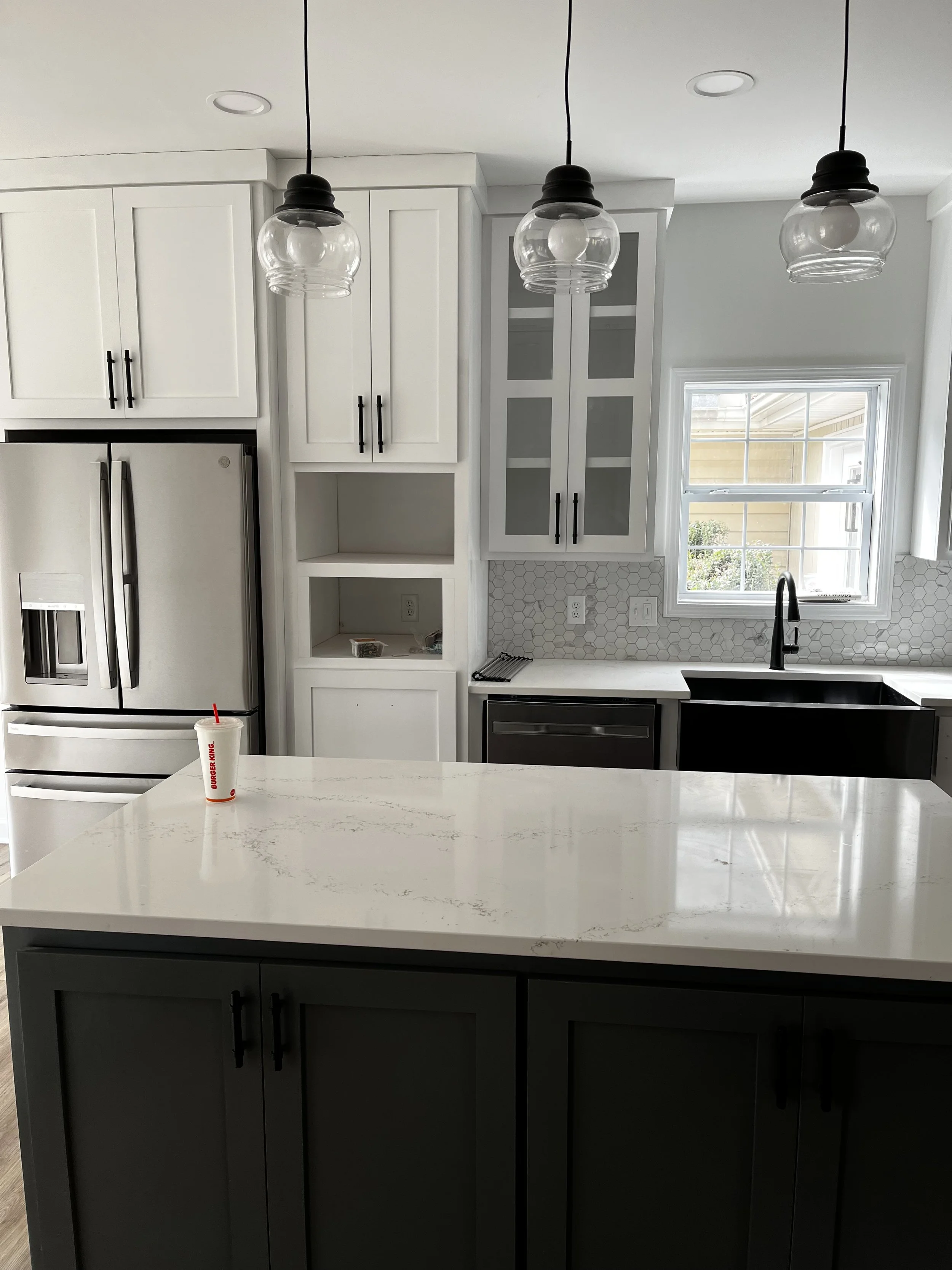 Modern kitchen with white cabinets, gray lower cabinets, a white marble island, stainless steel refrigerator, black fixtures, window above the sink, white hexagonal tile backsplash, and a drink cup on the island.