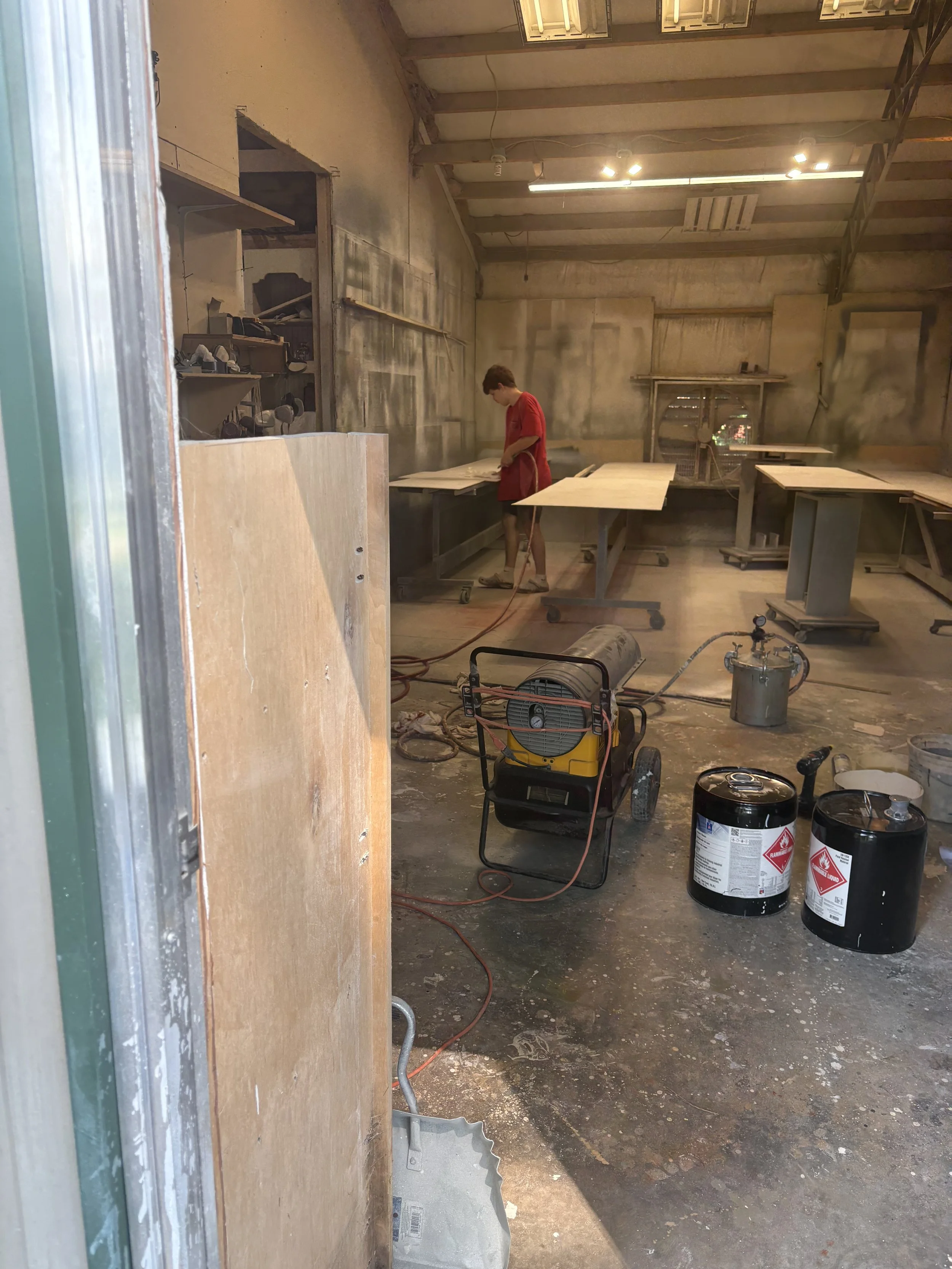 A person working in a woodworking or construction shop, surrounded by tools, wood, and industrial equipment, in an unfinished or renovation state.