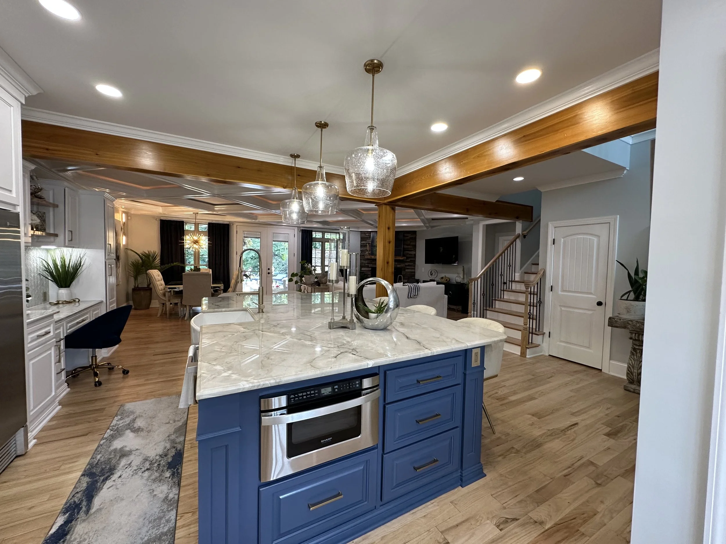 Open kitchen with white and blue cabinets, marble island, pendant lights, plants, hardwood floors, and a living area with a staircase and a TV in the background.