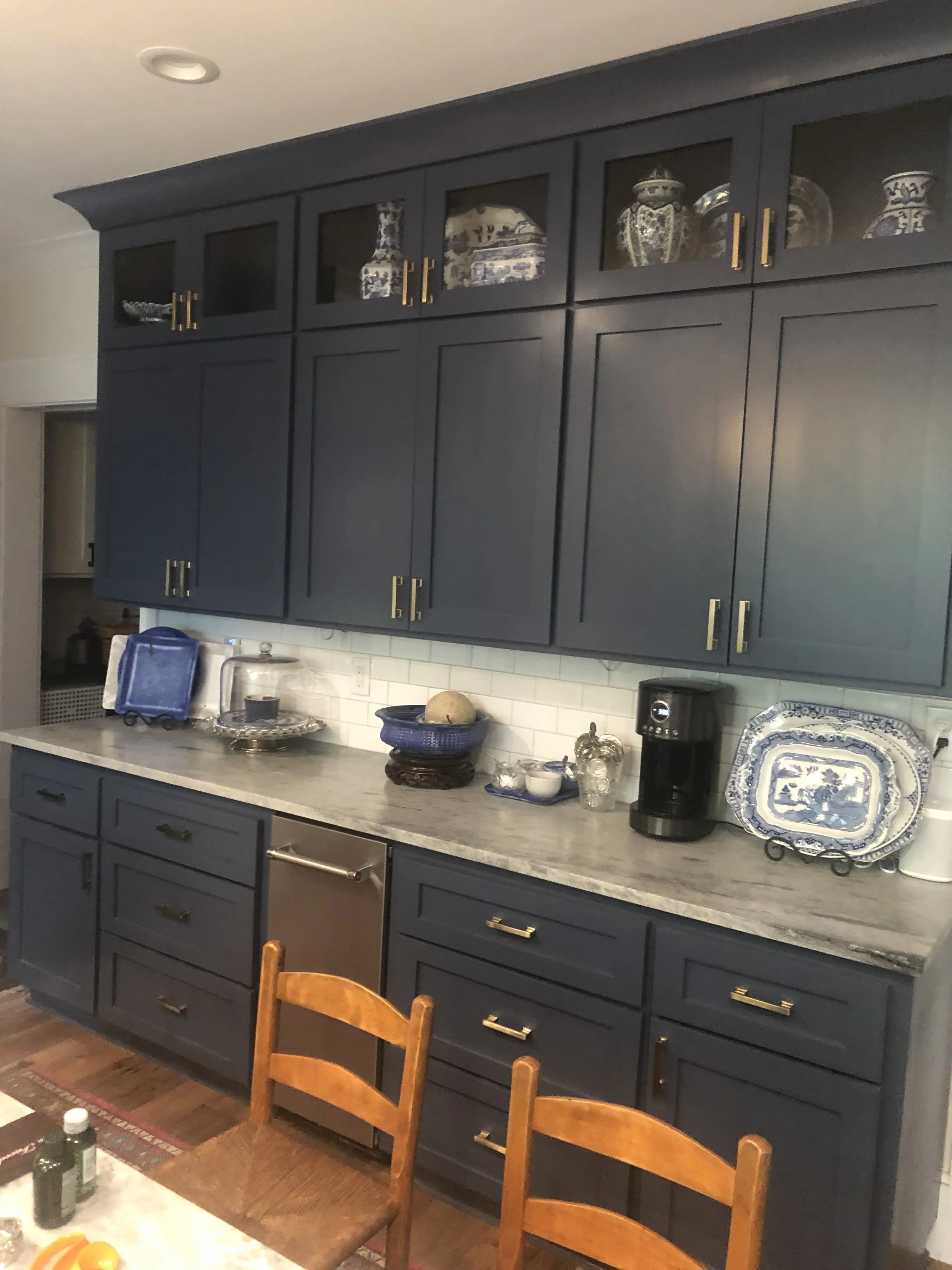 A kitchen with dark blue cabinets, some with glass-front doors displaying decorative vases. The countertop has dishes, bowls, and a coffee machine. Wooden chairs are partially visible in the foreground.