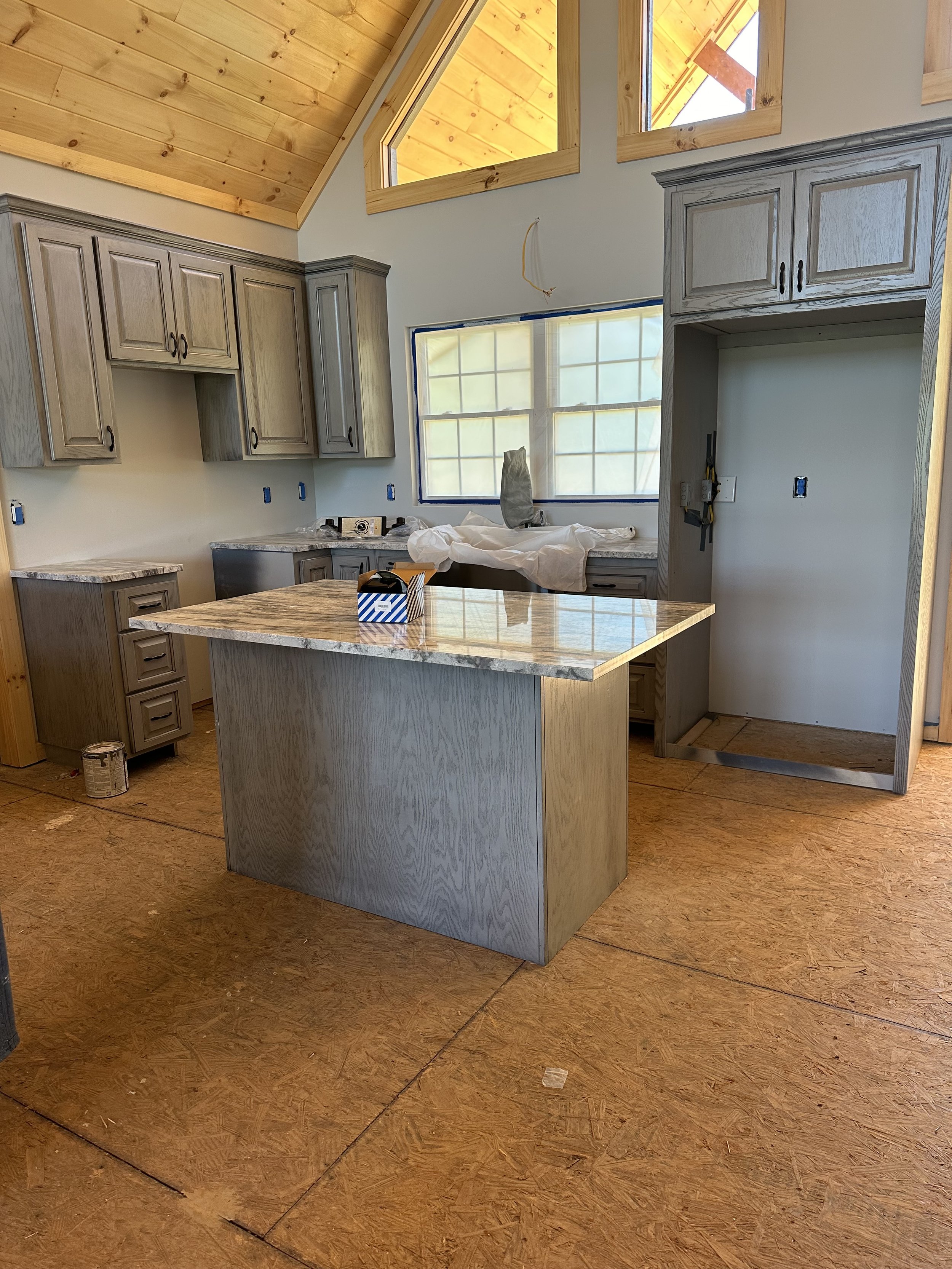 Interior of a kitchen under construction with unfinished cabinetry, a kitchen island, windows, and a wooden ceiling.