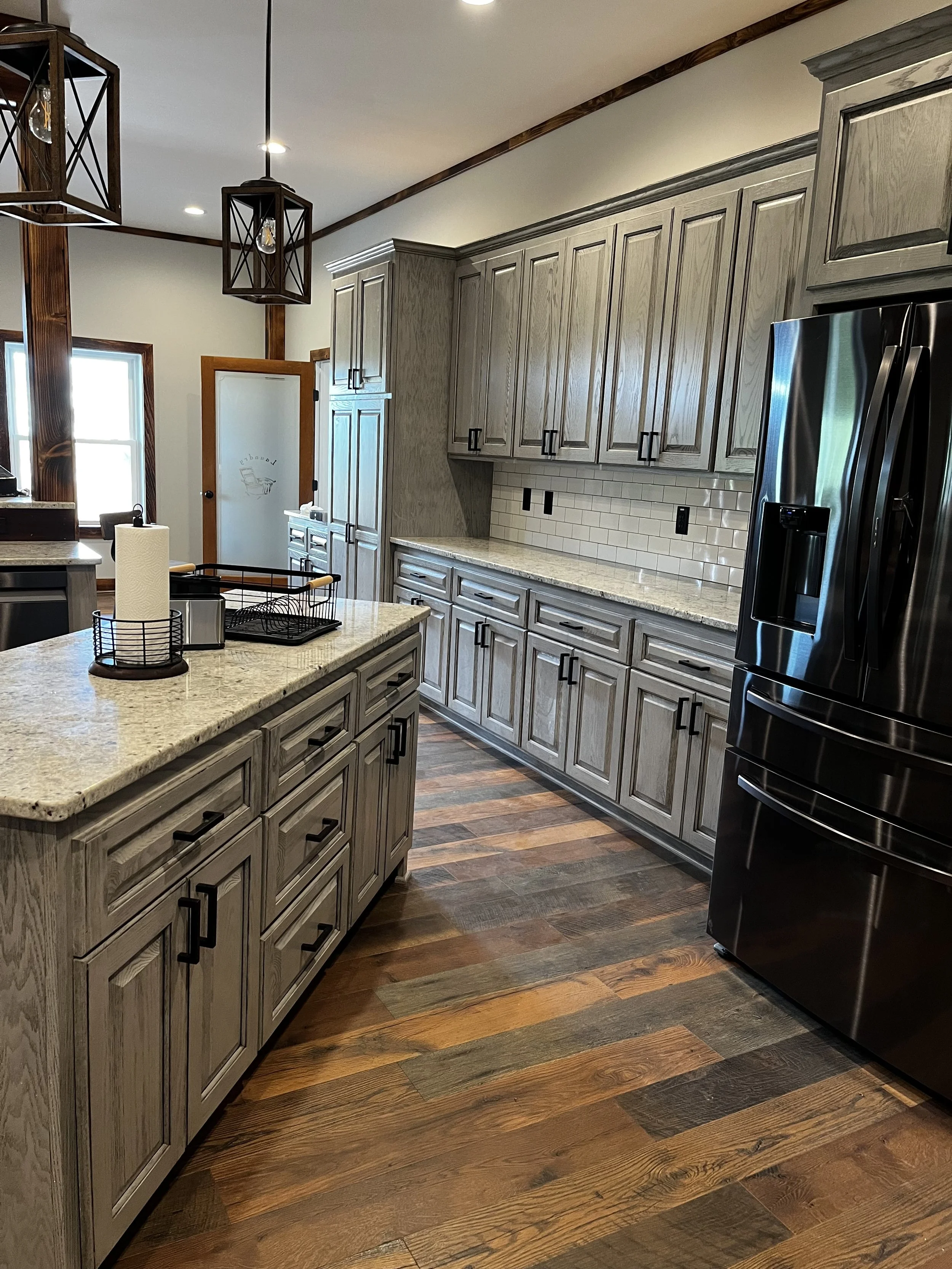 Modern kitchen with light gray wooden cabinets, granite countertops, and dark wood flooring. Stainless steel refrigerator, a lamp hanging from the ceiling, and a small island with a paper towel holder and wire baskets are visible.