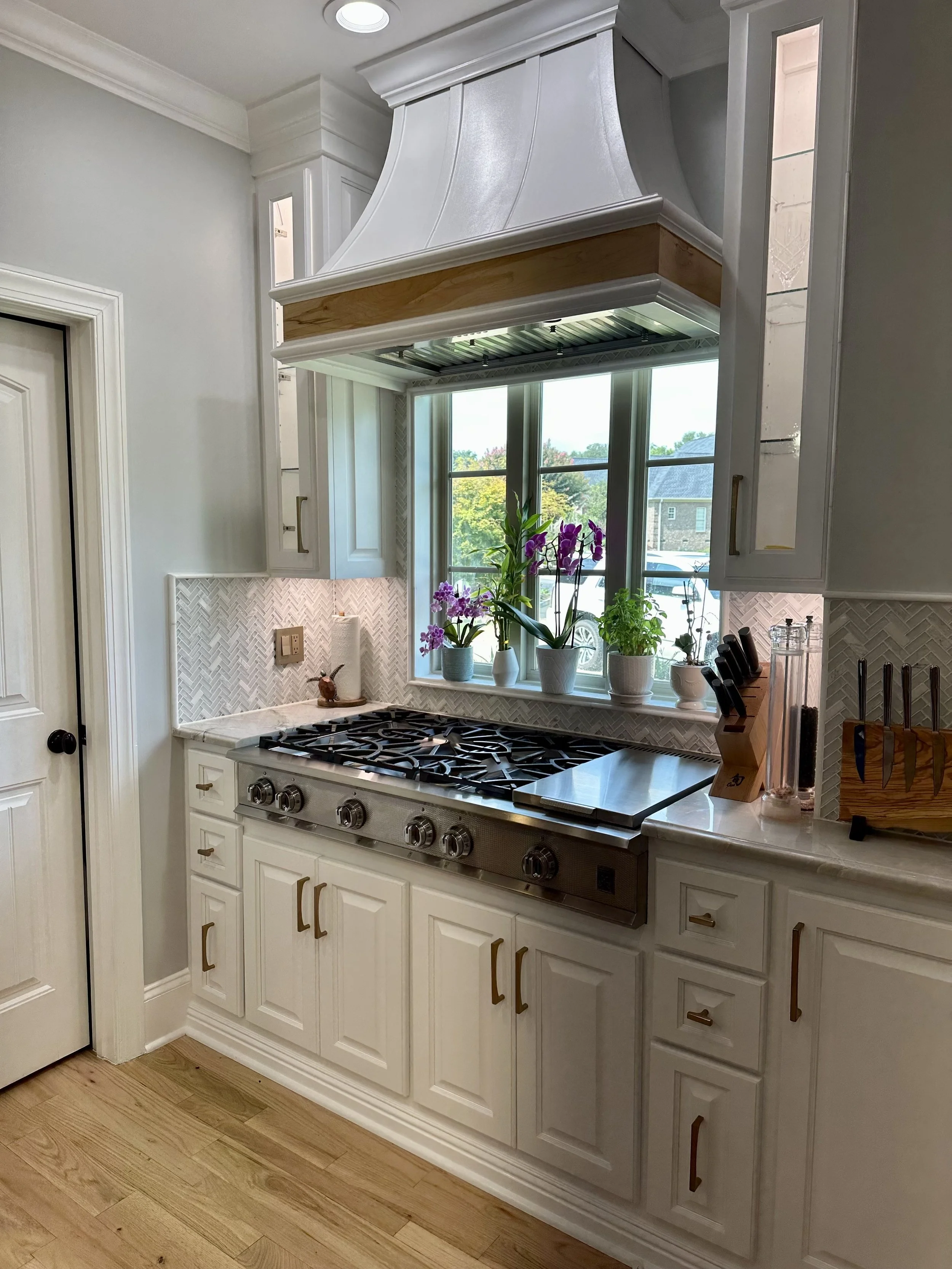 A kitchen with white cabinets, a gas stove, and a large window with plants on the sill. The kitchen has light-colored hardwood floors and a patterned backsplash.