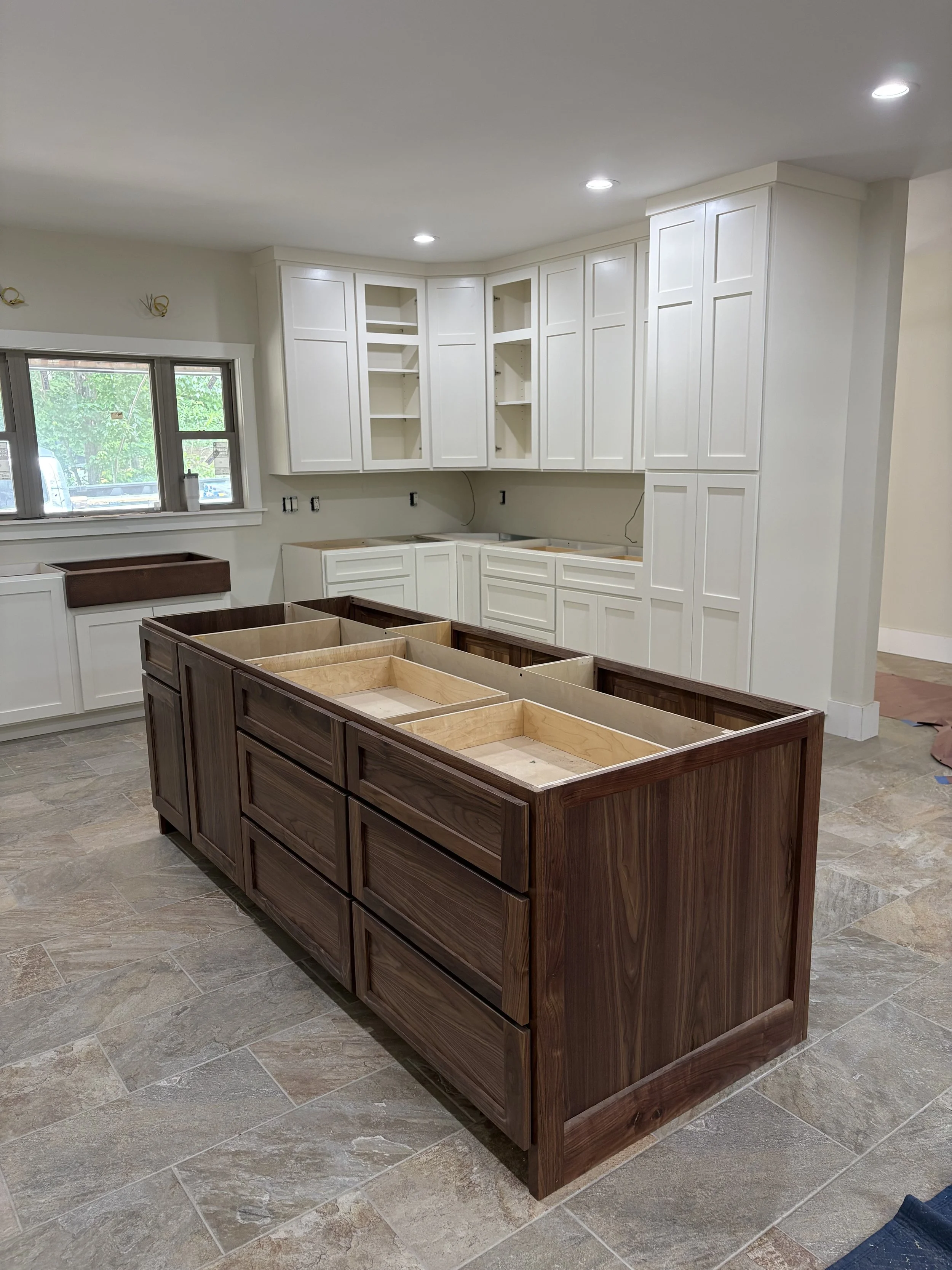 Kitchen under renovation with white cabinets and a wooden kitchen island in the center. The cabinets are partially installed, and the room has a large window over the sink area.