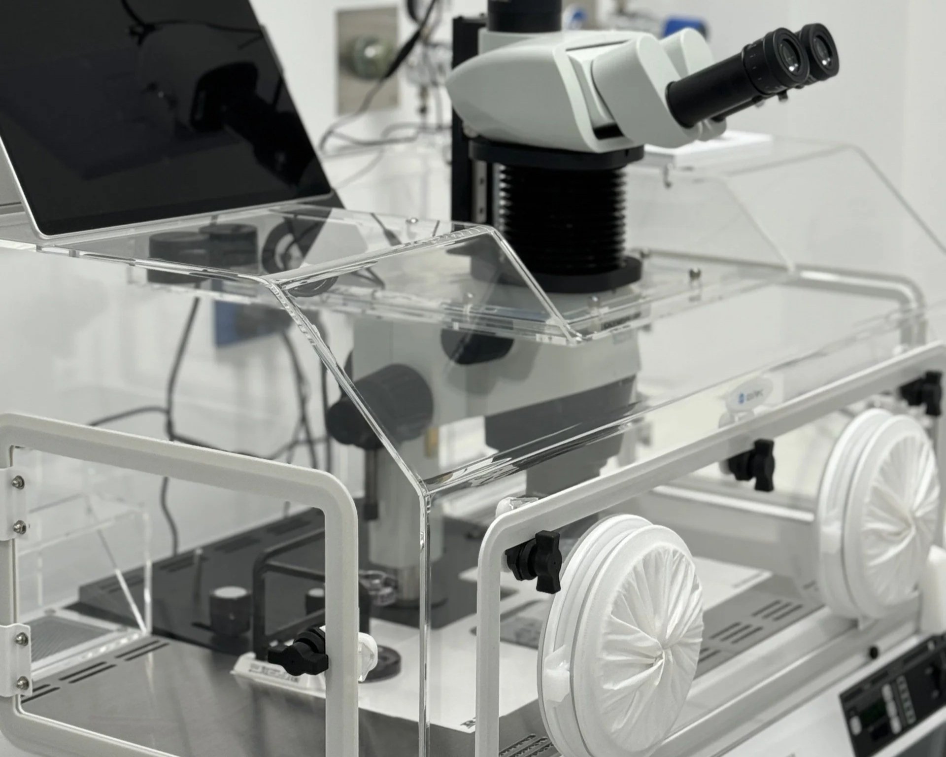 Laboratory microscope on a white stand with several white wheels, black knobs, and transparent barriers surrounding it.