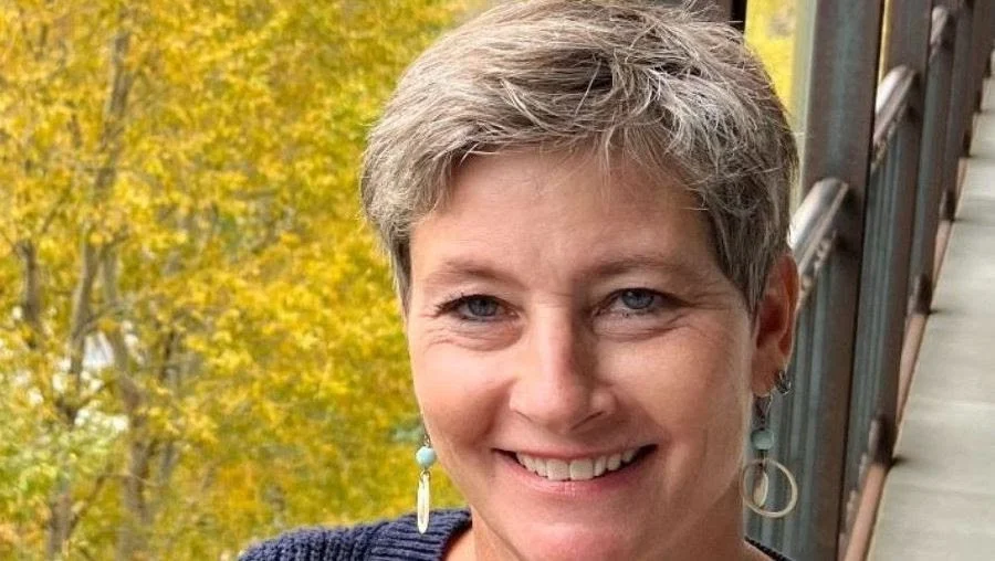 Close-up of a smiling woman with short gray hair, wearing earrings, outdoors in front of trees with yellow leaves.