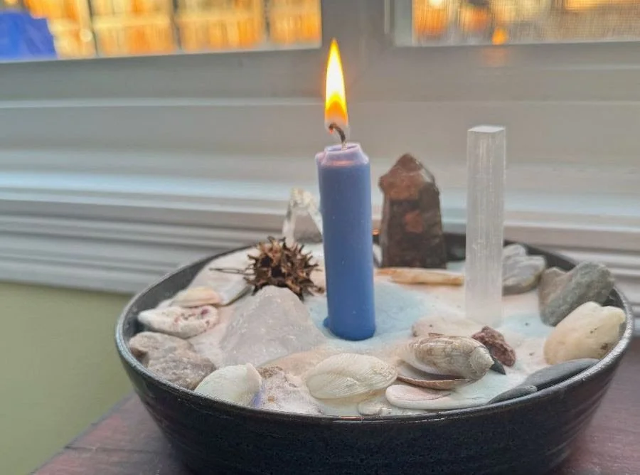 A black bowl filled with sand, seashells, rocks, and crystals, with a lit blue candle in the center, placed on a wooden surface near a window.
