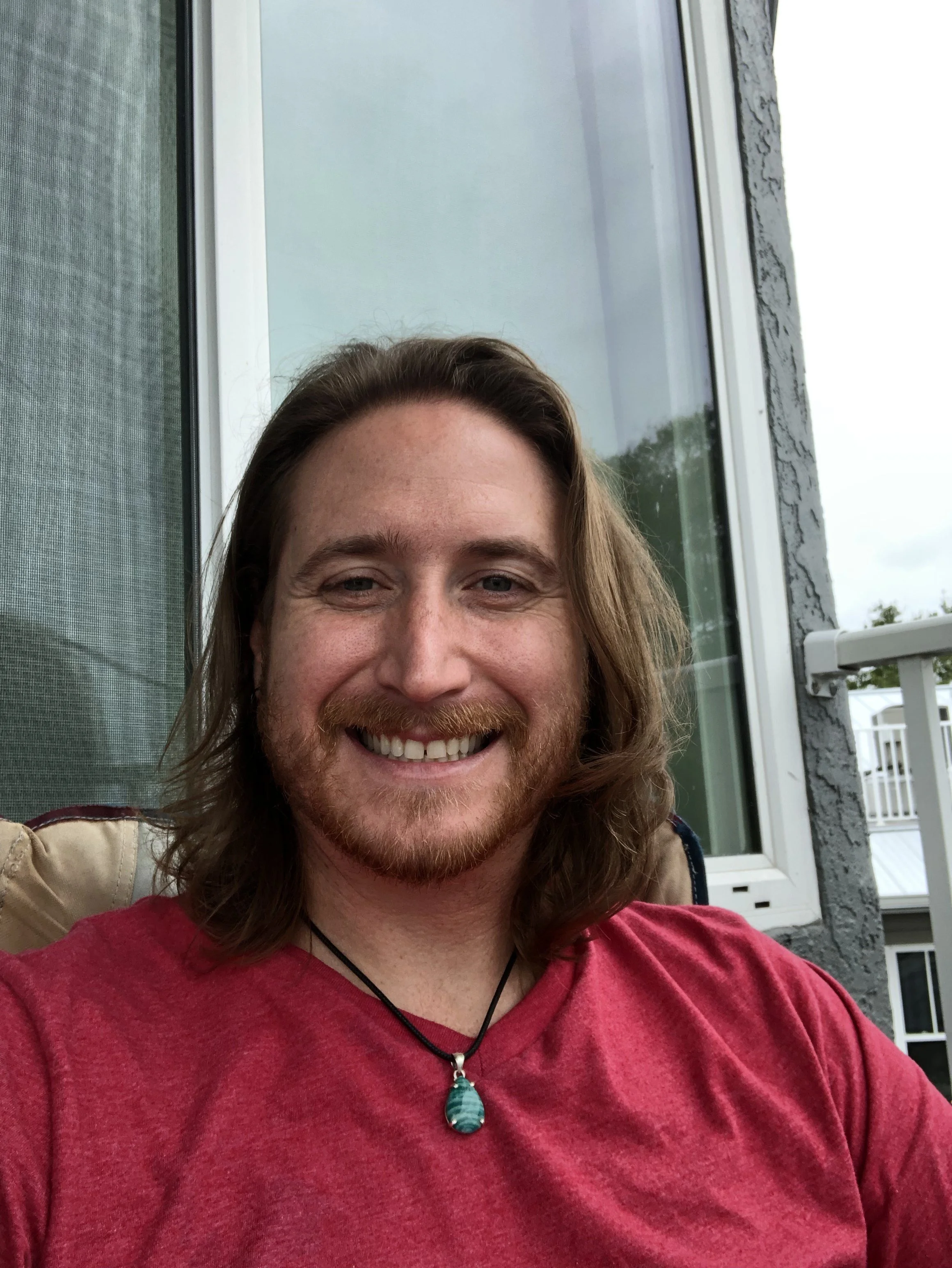 A smiling man with long hair and a beard, wearing a red shirt and a necklace with a large blue-green pendant. He is sitting outside on a patio or balcony, with a building and sky in the background.