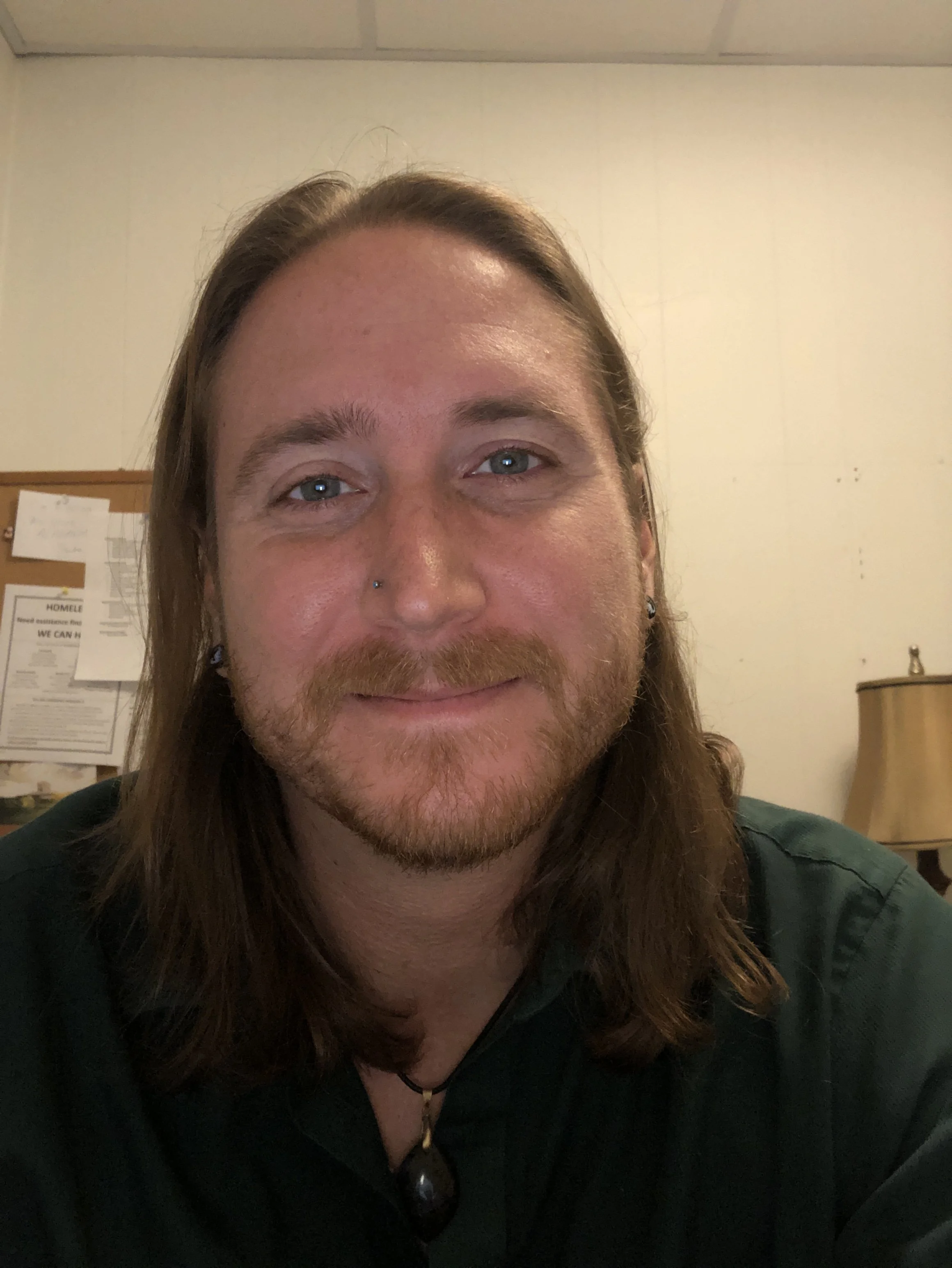 A close-up photo of a person with long brown hair, blue eyes, and a nose piercing, smiling slightly. The background shows a beige wall with papers pinned on a corkboard and a wooden lamp.