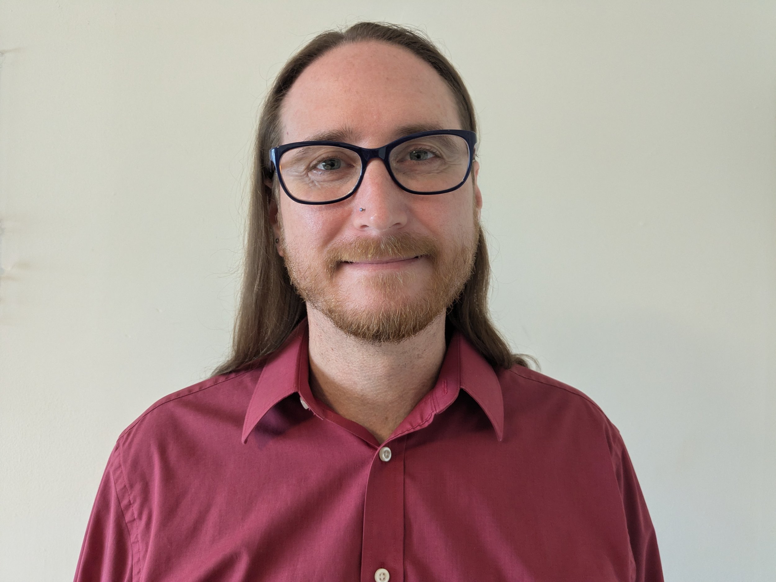 A man with long hair, wearing glasses, a nose piercing, and a maroon shirt standing against a plain light-colored wall.