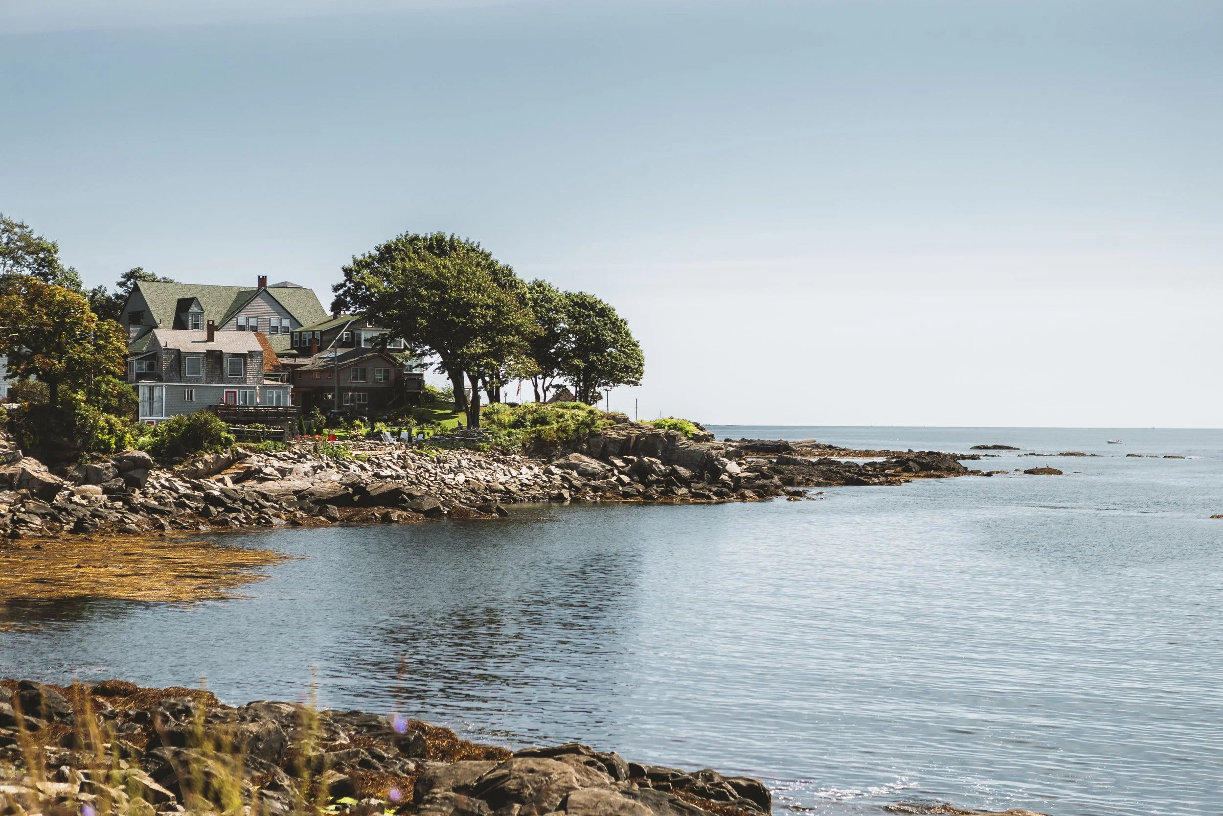 A coastal scene with houses on a rocky shoreline, trees, and calm water under a clear sky.